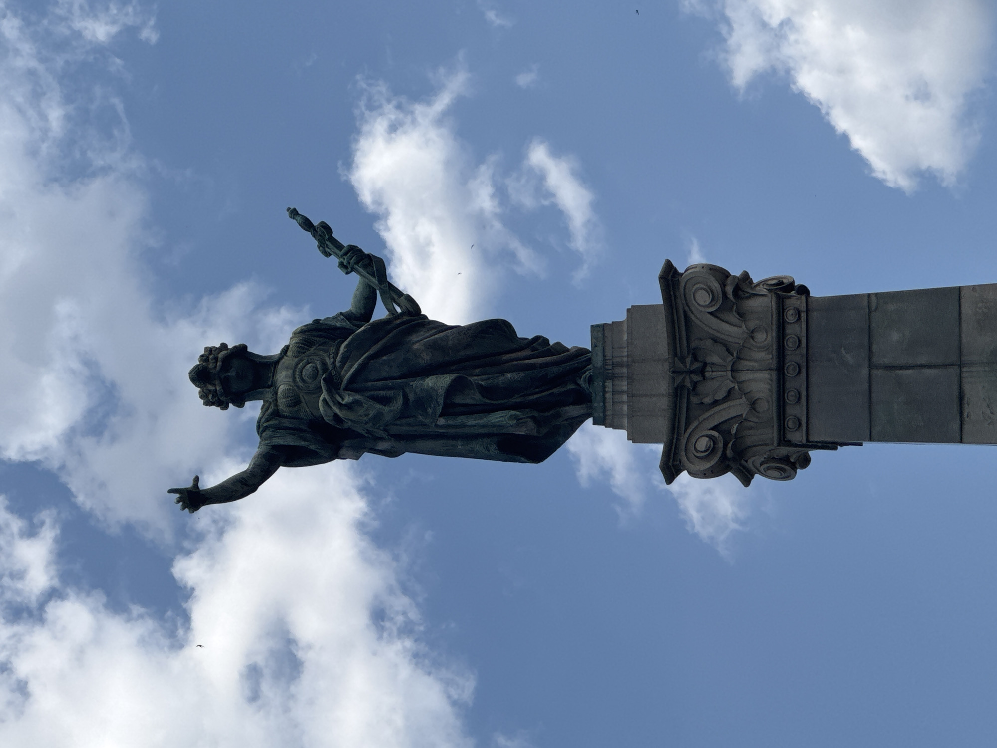 This media file features a statue of a woman in classical attire, holding a scepter in her right hand and reaching out with her left. The statue is made of dark metal, likely bronze, and displays intricate details in the drapery of her clothing and the design of the scepter. She appears to be standing atop a stone monument adorned with decorative carvings, including volutes and circular elements.
The perspective is from below, looking up at the statue against a bright blue sky filled with scattered white clouds. The lighting suggests it is daytime, likely mid-morning or afternoon, given the clarity of the sky and the absence of strong shadows that would indicate sunrise or sunset. A few birds are visible as small dark specks against the sky, adding a sense of movement and scale to the scene.
The overall impression is one of grandeur and permanence, with the statue serving as a focal point against the natural backdrop of the sky. The specific location is Ruse, Bulgaria, which might suggest the statue represents a historical figure, a symbol of the city, or an allegorical representation. There is no visible text in the image.