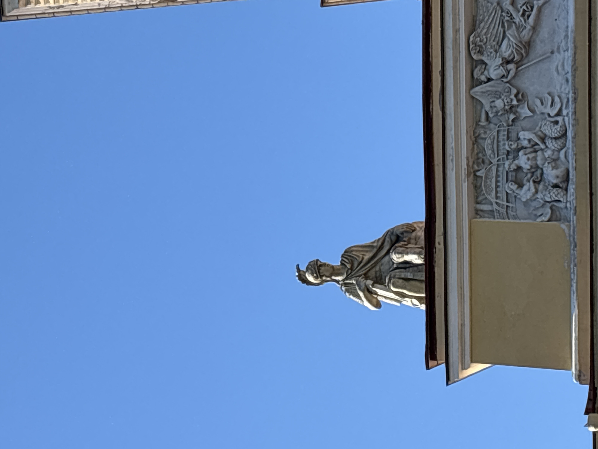 The image shows a stone statue of a person against a clear blue sky. The statue is depicted in profile, wearing a draped robe and a helmet. The person appears to be kneeling or seated, with one hand resting on an object that is not fully visible.
To the right of the statue, a section of a building is visible. This section features intricate stone carvings, possibly depicting mythological or historical scenes. The building appears to be old, with a light-colored facade and darker trim. The top of the frame also shows part of another building with a similar architectural style, suggesting a grand or historical setting.
The clear blue sky indicates it is daytime, likely with good weather. The angle of the shot, looking upwards, emphasizes the scale of the statue and the building. The overall impression is one of architectural grandeur and artistic detail. While specific identification of the statue or building is not possible from the image alone, the style of the architecture and sculpture, combined with the given location context of Saint Petersburg, Russia, suggests it could be a historical monument or part of a significant landmark in the city. No visible text is present in the image.