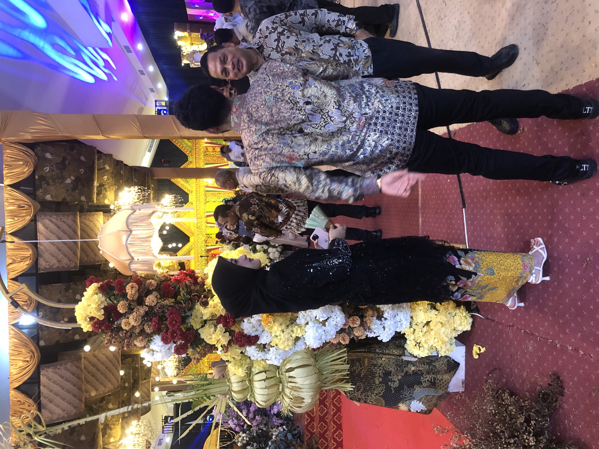 The image depicts an indoor event, likely a wedding reception, held at the Kuta Raja Hotel in Lhokseumawe, Indonesia. A woman in a black hijab and a sequined dress stands to the left, holding a light pink object. She is positioned near a large floral arrangement and a decorative tower made of dried leaves. To her right, two men in batik shirts are engaged in conversation. The man in the foreground is facing the camera, smiling, while the man in the background has his back to the camera. The environment is decorated with ornate drapes, chandeliers, and colorful lighting. The floor is covered with a red carpet with a patterned design. The overall atmosphere suggests a formal celebration.