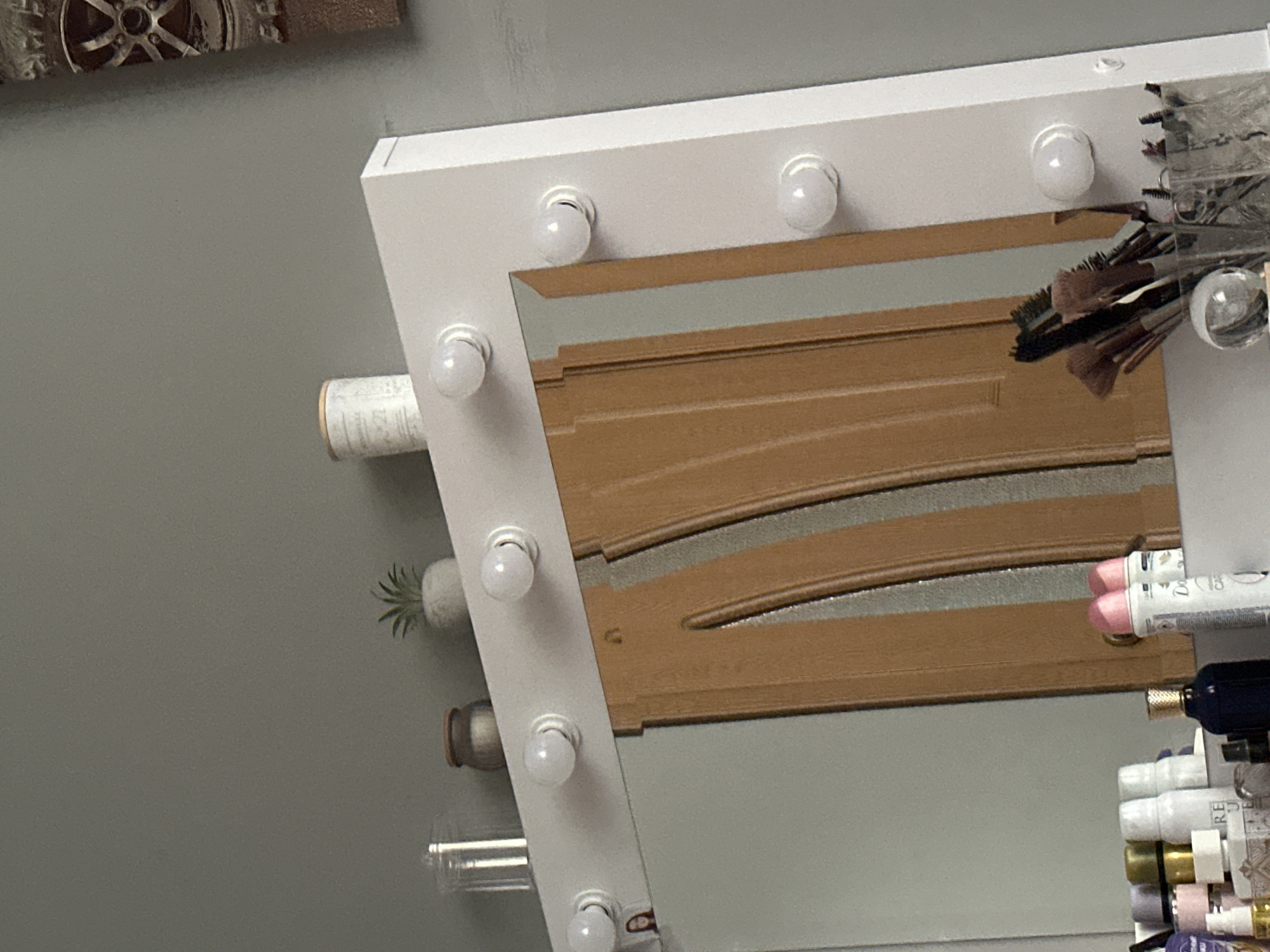 An indoor, eye-level shot shows the right side of a white vanity mirror with illuminated bulbs and a reflection of a wooden door. The mirror is framed in white and features several round, white bulbs evenly spaced along its top and right side. A small, framed picture of a woman is visible on the left side of the mirror. The reflection in the mirror shows a light brown wooden door with vertical paneling. On top of the mirror frame, to the left, are two small decorative items: a clear glass cylinder and a small, grey pot with a green succulent plant. To the right of the succulent is a cylindrical white container with a textured surface. In the bottom section of the image, various makeup items are visible, including makeup brushes in a clear container and several bottles and tubes of cosmetics. The background is a plain, grey wall.
