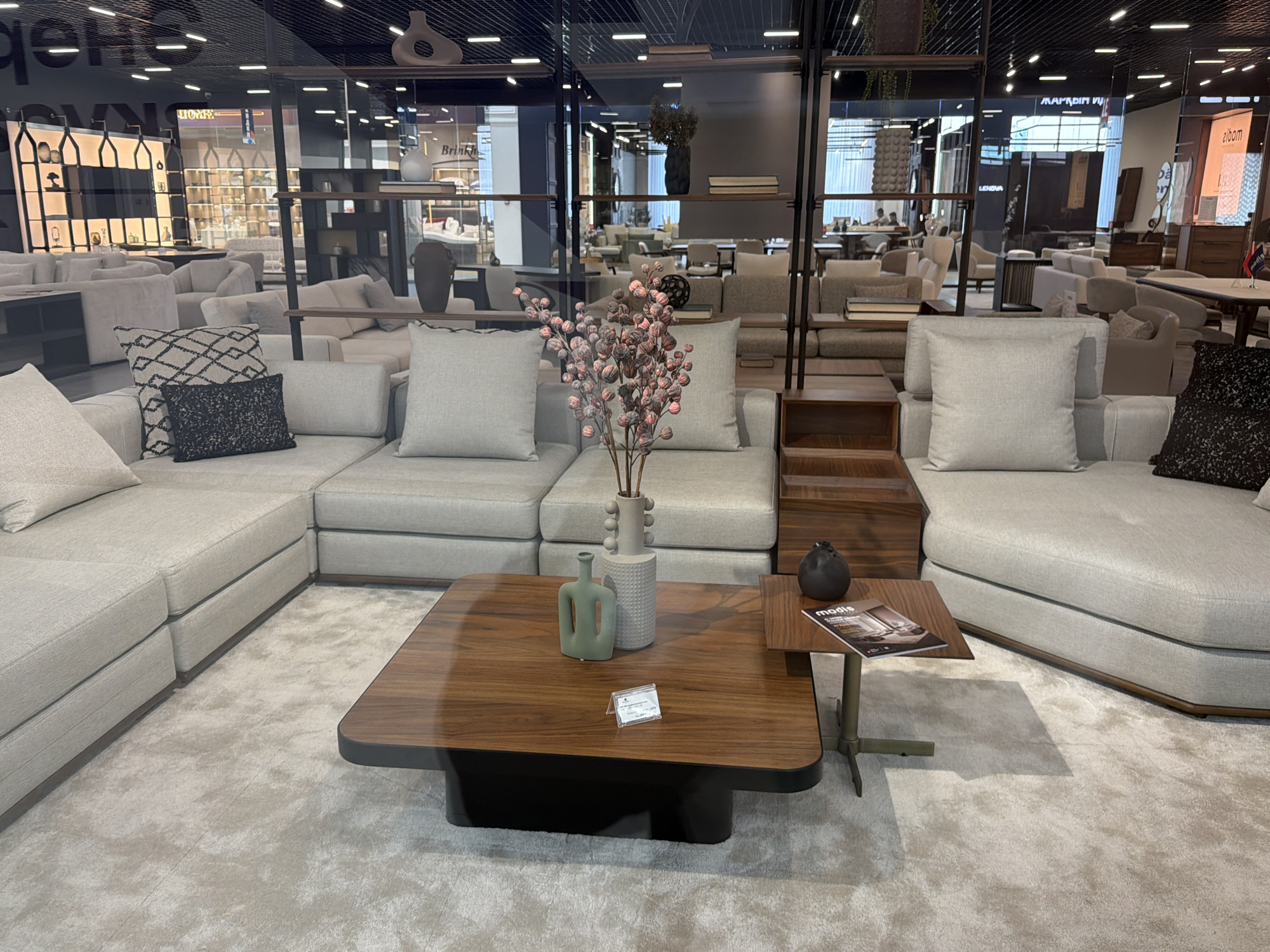 The image depicts a well-lit showroom interior, likely a furniture store, with a focus on a modern, modular sectional sofa in a neutral, light gray fabric. The sofa is arranged in a large L-shape, with multiple cushions and a low-profile design. In the foreground, a dark wood coffee table with rounded edges sits on a plush, light-colored rug. On the coffee table, there are two decorative vases: one tall, cylindrical, light gray vase with a textured pattern, and a smaller, mint green vase with an abstract, geometric shape. Next to the coffee table, a smaller side table holds a black spherical object and a magazine with the word "modis" visible on its cover.
Behind the sofa, a dark wooden shelf unit with multiple levels is visible, displaying various decorative items, including vases and books, as well as some greenery hanging from above. The background shows more furniture arrangements, including additional sofas and chairs, suggesting a larger showroom space. Reflections in the glass walls of the showroom reveal more of the store's interior, including displays of other furniture and lighting fixtures. The overall impression is one of a contemporary and sophisticated retail environment. There are no people clearly visible in the immediate foreground or mid-ground. The lighting is bright and even, indicative of an indoor setting during business hours. The location context of Astana, Kazakhstan is not directly evident from visual cues within the image itself, but provides external information about the setting.