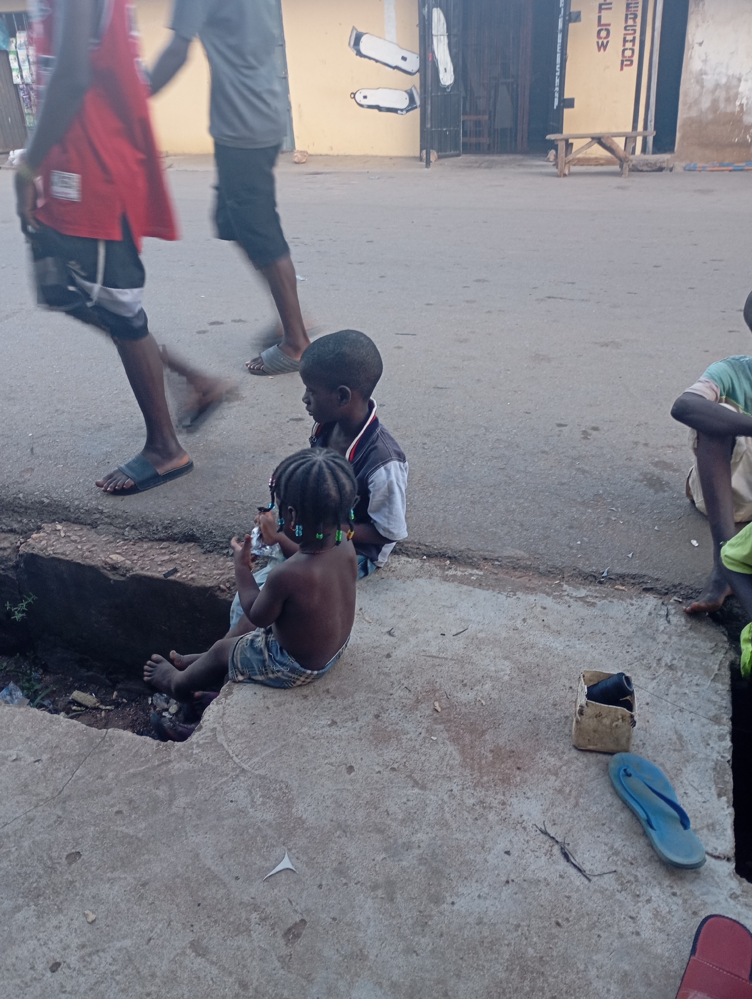 In the city of Unknown, Unknown, three children are visible in the foreground. Two children, a boy and a girl, are sitting on a concrete ledge. The girl is facing away from the camera, wearing a plaid skirt and holding a clear plastic bag. The boy is facing the girl, wearing a collared shirt. A third child, partially visible, is sitting to the right. In the background, two adults are walking. One is wearing a red sleeveless shirt and dark shorts. The other is wearing a grey t-shirt and dark shorts. The environment appears to be a street, with a building in the background displaying text. A wooden bench is placed in front of the building. Scattered objects on the ground include a blue flip-flop, a small cardboard box with a spool of black thread, and other debris.
