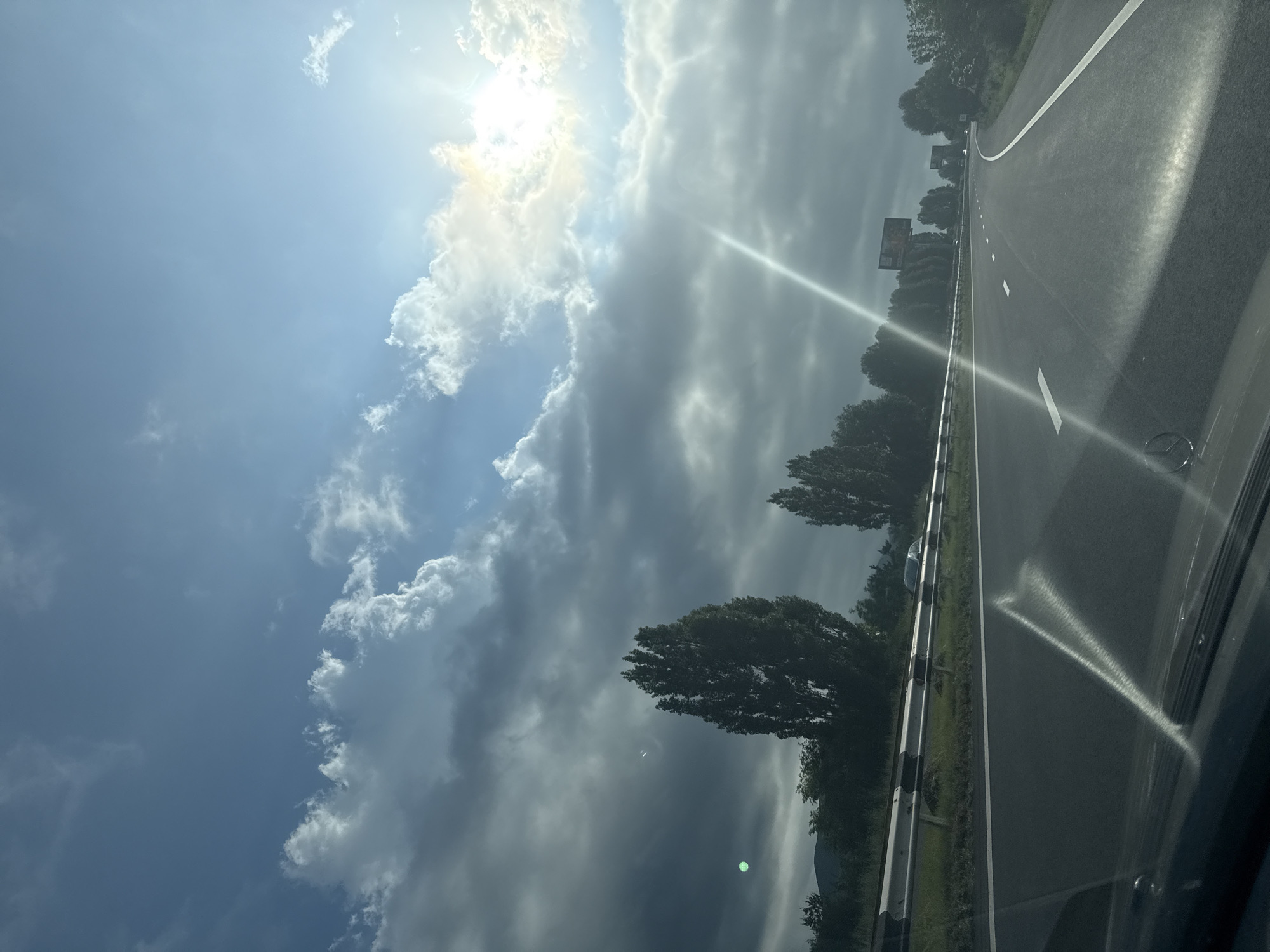 A car is driving on a highway with a guardrail on the left and trees on the right. The sky is partly cloudy with the sun shining through. The sun's rays create a lens flare across the windshield. The scene appears to be during daylight hours. The location is identified as the city of Gagarin in Armenia.
