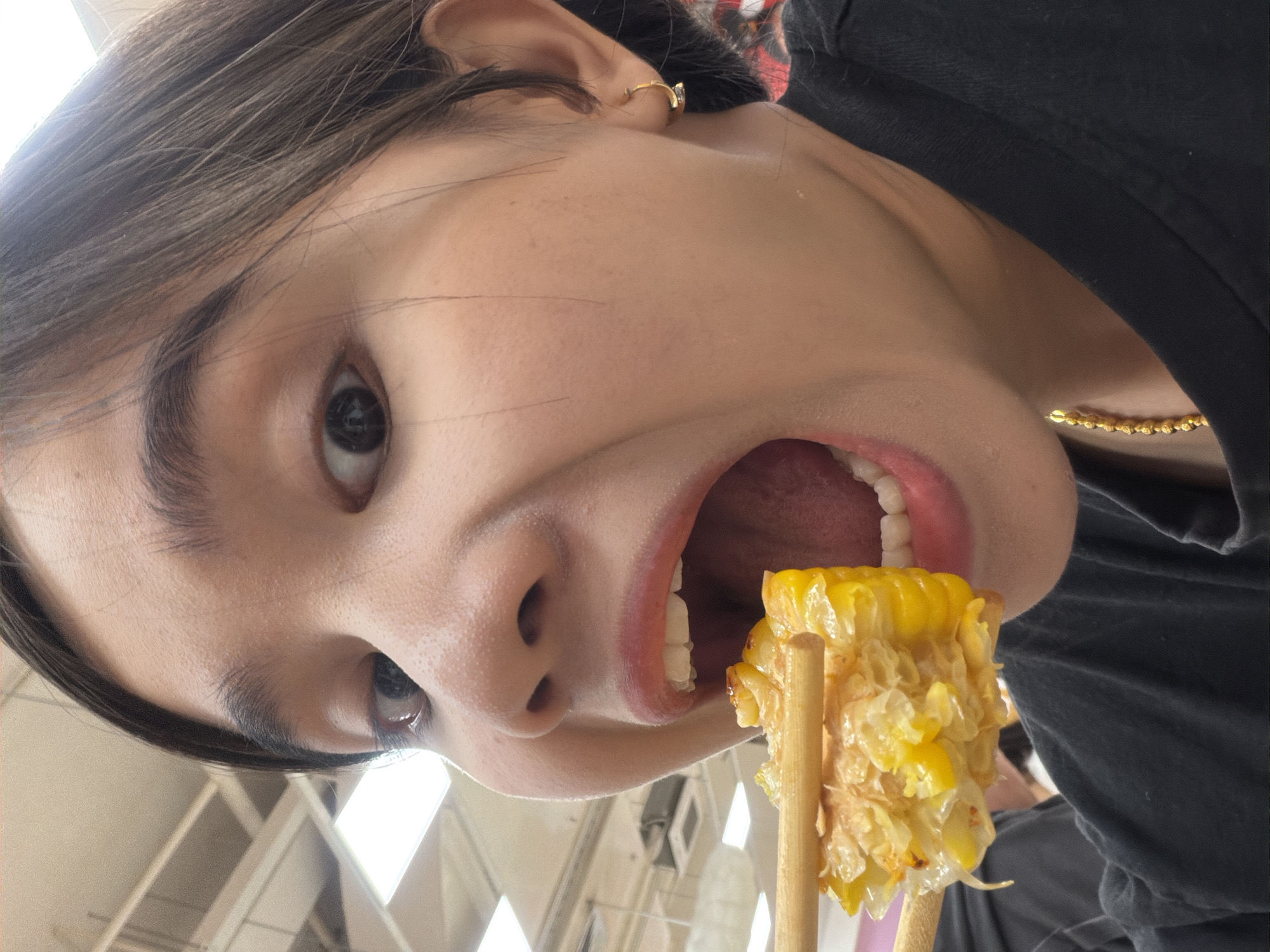 A young person is seen in a close-up, angled shot, holding a partially eaten corn on the cob impaled on two wooden sticks, positioned directly in front of their open mouth. The person has dark hair, light skin, and is wearing a black top. A small gold hoop earring is visible in their left ear, and a gold necklace is partially visible around their neck. The background suggests an indoor environment with bright, artificial lighting. The setting is claimed to be in Đà Nẵng, Vietnam, but visual confirmation is not evident within the frame.