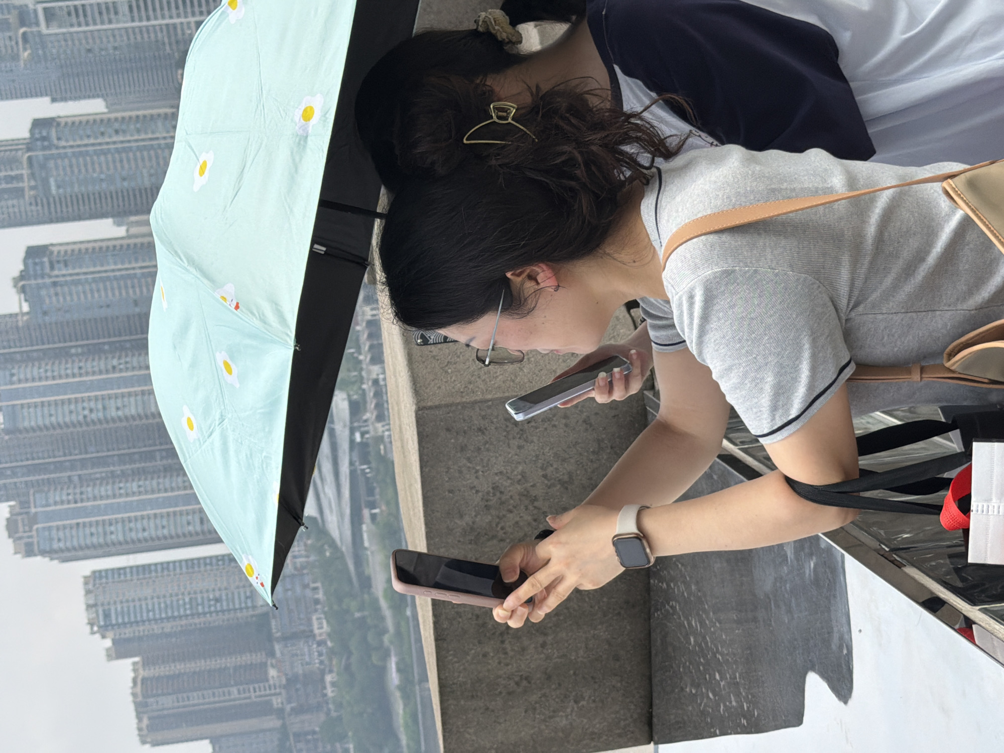 Two women stand under a light blue umbrella decorated with yellow and white daisy-like patterns, facing away from the camera. They are positioned on a concrete ledge overlooking a cityscape with numerous tall, modern buildings, suggesting an urban environment like the Yuzhong District in China. The woman on the left, wearing glasses and a grey t-shirt, holds a smartphone in her right hand, seemingly taking a photo or recording a video. She also wears a light-colored smartwatch on her left wrist. Her hair is pulled back, and she is carrying a black tote bag with a red item visible inside, along with a tan crossbody bag slung over her shoulder. The woman on the right, whose back is mostly to the camera, wears a dark blue and white top and has her dark hair tied up with a scrunchie. She is holding a smartphone in her right hand. The sky is overcast, and the ground in front of the women is wet and reflective, indicating recent rain.