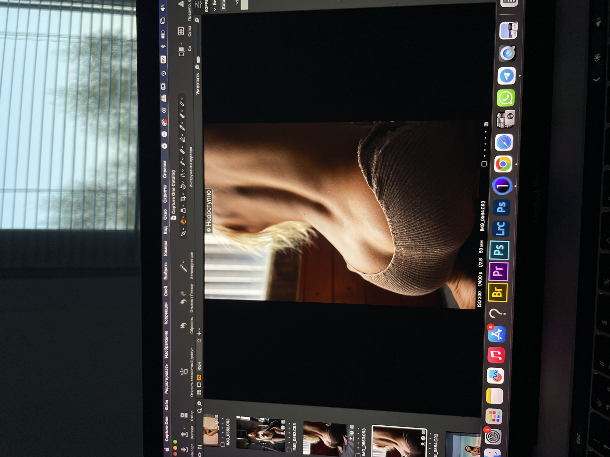 The image is a photo of a MacBook Pro screen displaying the Capture One software. Within Capture One, a photograph of a woman's shoulder and neck is visible. The software interface shows various tools, menus (File, Edit, Image, Corrections, Layer, Select, Camera, View), and import/export options. The application’s window also contains thumbnails of other images from the same photoshoot. The macOS dock on the right side of the screen contains various application icons, including Safari, Google Chrome, Capture One (labeled "1"), Adobe Photoshop, Adobe Premiere Pro, and others. The physical environment shows a window with blinds and a laptop keyboard. The location in Kazan, Russia cannot be determined from the information provided.