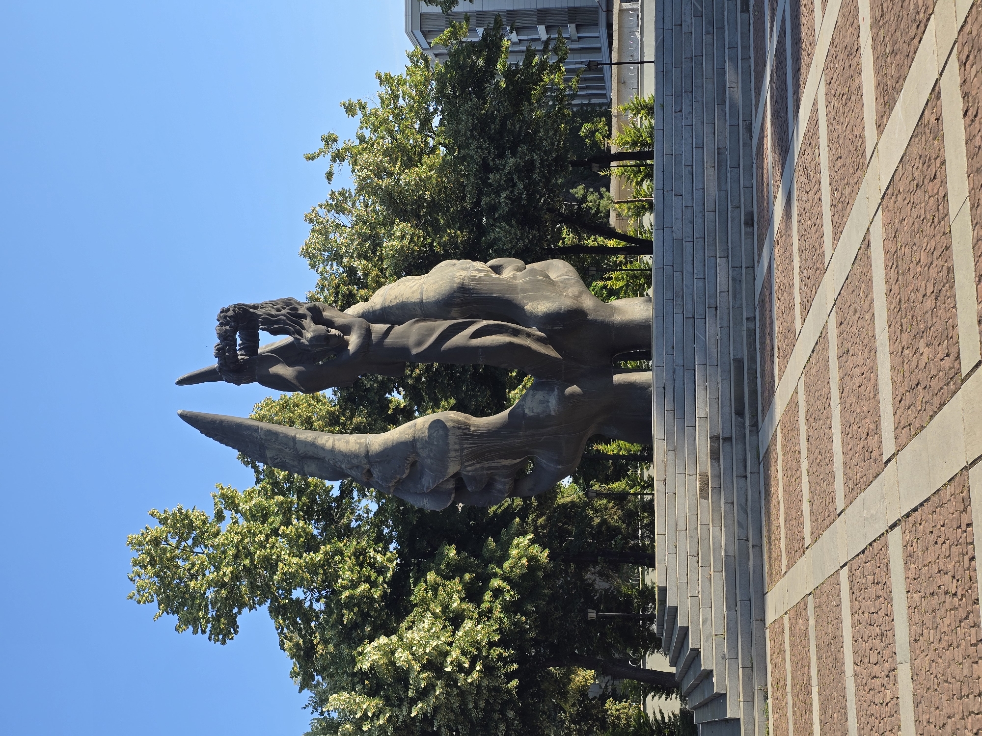 A monumental sculpture stands in an open plaza, bathed in sunlight under a clear blue sky. The sculpture is composed of two large, dark, stylized hands forming a concave shape, supporting a human figure crowned with a wreath. The figure appears to be holding the wreath aloft. Behind the sculpture, a wide set of stone steps leads up to a platform, and beyond that, lush green trees form a dense canopy. In the distance, a modern building with a geometric facade is visible. The plaza floor is paved with reddish-brown cobblestones arranged in a grid pattern with lighter stone borders. This is the Monument to the Soviet Army in Plovdiv, Bulgaria, also known as Alyosha.