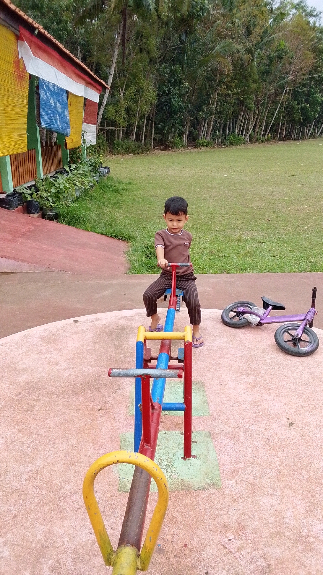 A young boy is sitting on a seesaw in a playground in Cintaratu, Indonesia. He is wearing a brown shirt and striped pants, and his feet are bare. The seesaw is red, blue, and yellow. To the right of the seesaw, a purple bicycle lies on the ground. The playground is situated on a grassy field with trees in the background. A building with yellow and red banners is visible on the left side of the image.