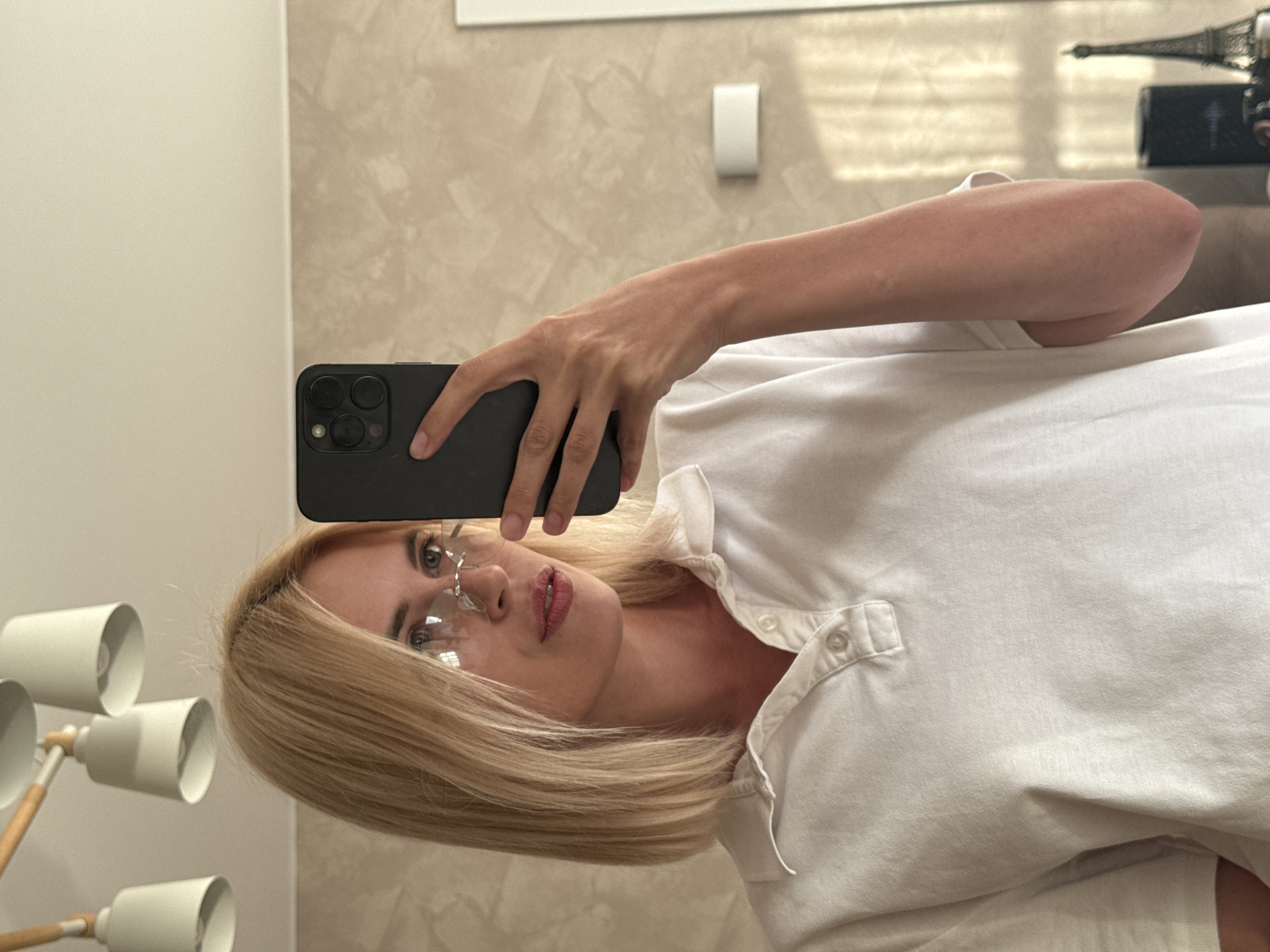 A young woman with blonde bobbed hair and striking cat-eye glasses is taking a selfie in a mirror. She is wearing a white polo shirt and her left arm, holding a dark-colored smartphone, is extended upwards to capture the shot. The background appears to be an interior space with a textured wall and decorative elements like a modern chandelier and a miniature Eiffel Tower on a shelf. The lighting suggests it could be daytime with natural light filtering in, possibly from a window. The location is not specified in the image or the provided context.