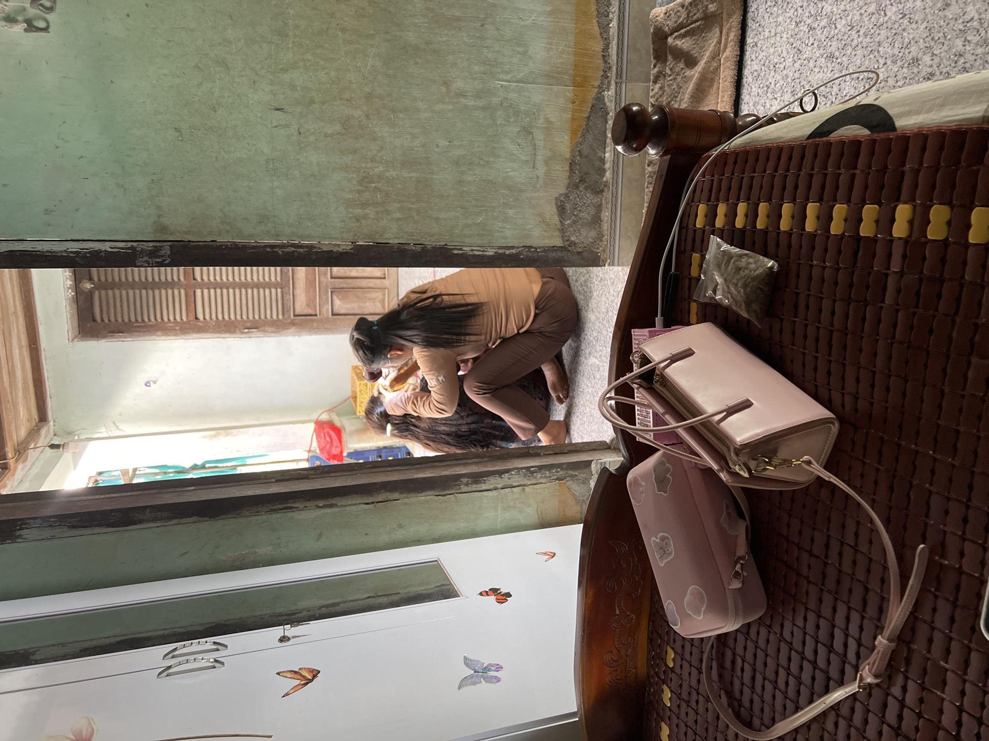 The image captures an indoor scene, looking into an adjacent room or hallway within a home in Bình An, Vietnam. In the immediate foreground, on the right, a patterned brown mat, likely made of bamboo and possibly covering a bed or bench, holds two pink bags: a larger shoulder bag and a smaller pouch decorated with cartoon clouds and animals. A small clear plastic bag containing a granular substance and a white charging cable are also present on the mat.
Further into the scene, through an open doorway, a person with long dark hair, wearing a brown top and matching pants, is seen from behind. They are bending down close to the floor, with their bare feet visible, appearing to be focused on or interacting with something or someone mostly obscured in front of them.
The background reveals more details of the interior. To the left, a white wardrobe or cabinet adorned with decorative butterfly stickers and silver handles is visible. Directly opposite, further in the background, a wooden window with slatted shutters allows natural light into the space. A red plastic item, possibly a container, can be vaguely seen in the distance. The walls appear somewhat aged, consistent with a residential setting. The scene is well-lit, indicating it is daytime. No discernible text is visible in the image.