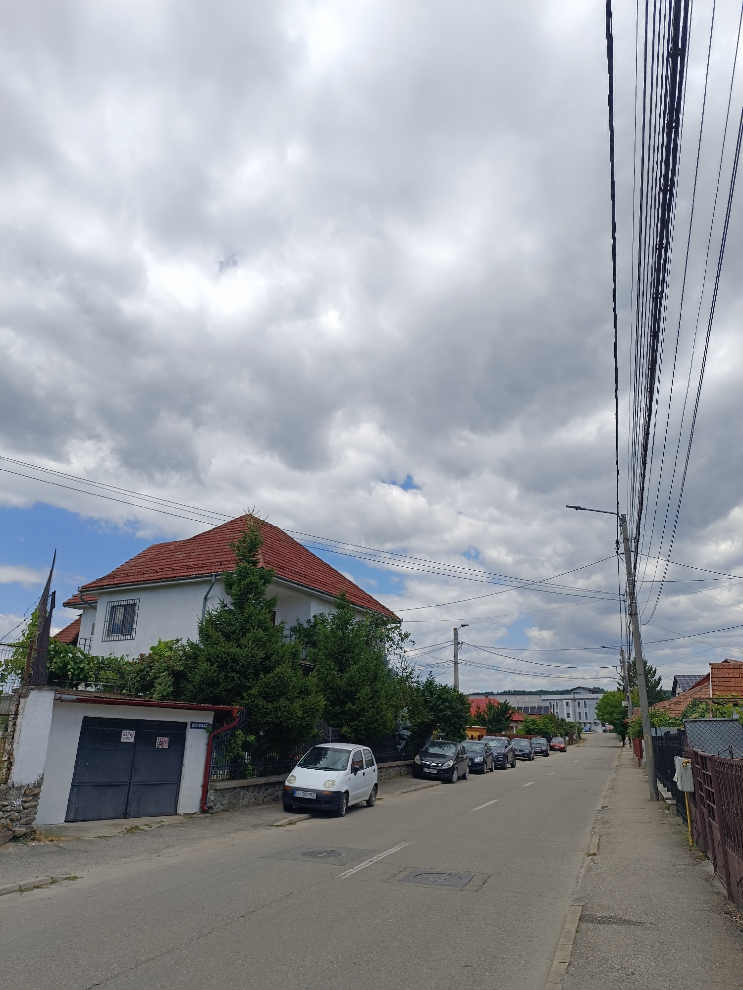 The image depicts a street scene in Târgu Jiu, Romania, under a cloudy sky. The street is paved and has a curb. On the left side of the street, there is a white house with a red tiled roof, bordered by lush green trees and bushes. A white garage with dark gray doors is visible in front of the house. Adjacent to the garage, a small white car is parked. Further down the street, several other cars are parked along the side. On the right side of the street, a sidewalk runs alongside a brown metal fence. A utility pole with numerous overhead wires stands tall. In the distance, more houses and buildings can be seen, some with red tiled roofs and solar panels. The overall impression is a residential street in a town or city.