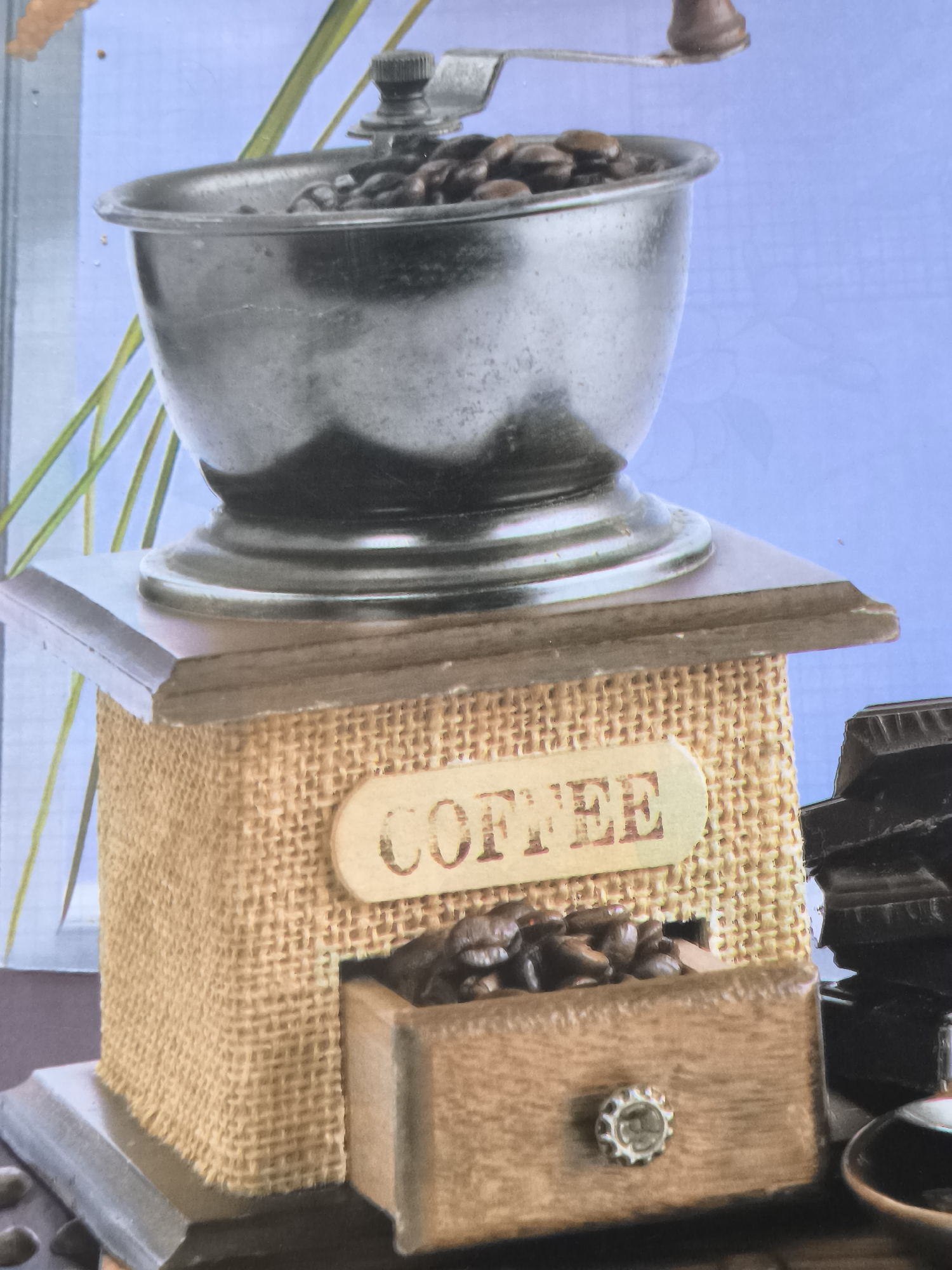 This is a close-up, eye-level, medium shot of a decorative coffee grinder and some dark chocolate bars. The coffee grinder has a metal hopper filled with coffee beans, a wooden crank on top, and a burlap-covered body. Below the burlap, there is a small wooden drawer containing more coffee beans, with a decorative knob on the front. A sign with the word "COFFEE" is attached to the burlap body. To the right of the grinder, a stack of dark chocolate bars is visible. The background is a light blue with a subtle floral pattern and some green plant stalks. The scene is static and appears to be a still life arrangement. There are no people or animals present. The location is not specified to be in Dobrogea Veche, Moldova.