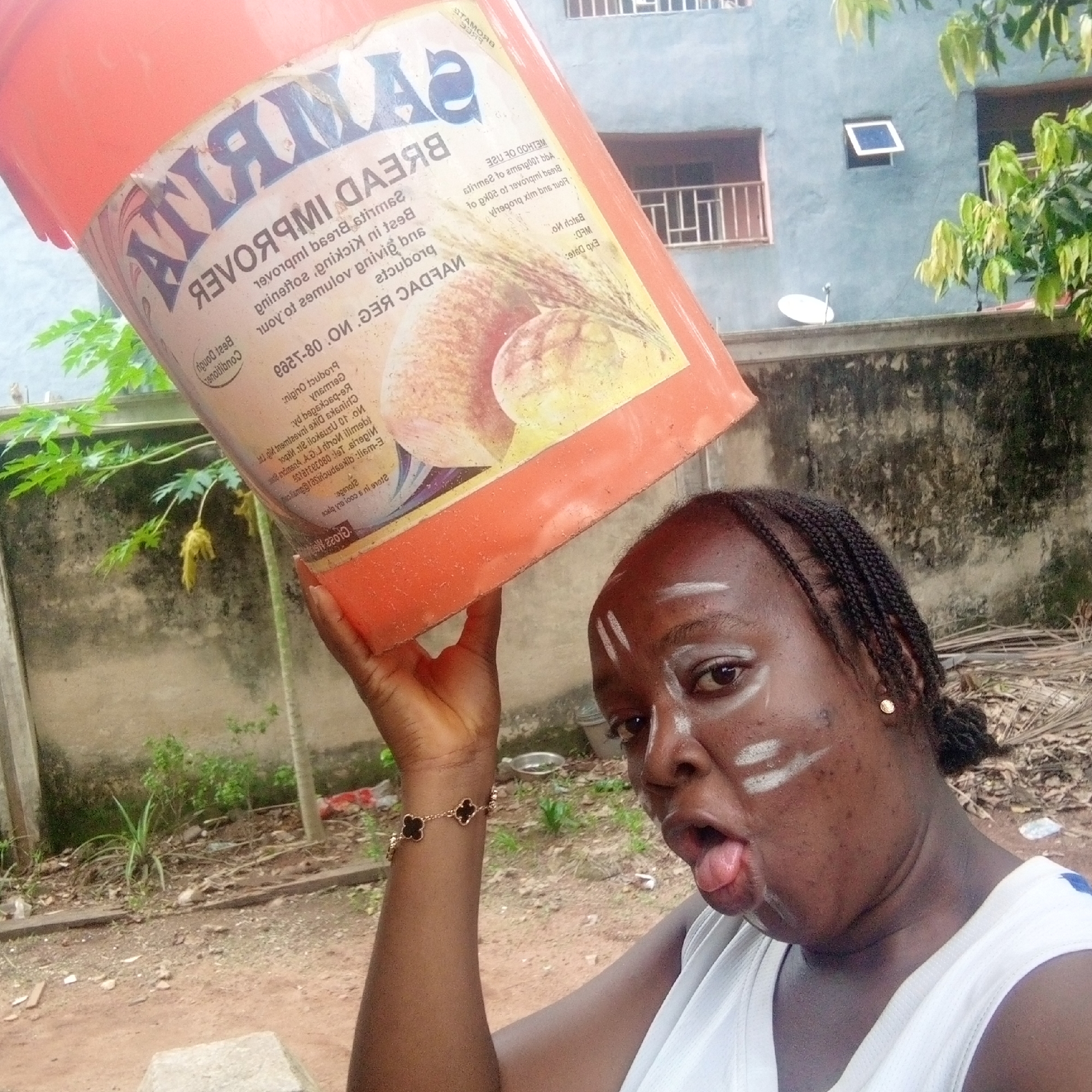 A woman is standing outdoors holding a large orange bucket over her head. The bucket is labeled "ATIAMAS BREAD IMPROVER." The woman is wearing a white tank top and has tribal-like white paint on her face. Her tongue is sticking out. She has braided hair and wears a gold bracelet and earring. The background features a yard, a fence, a papaya tree, and a two-story building. The bucket's label also mentions "Nigeria" on it. Given the label and references on the bucket, coupled with tribal makeup elements, it is highly likely the image was taken in Nigeria; Ogidi is not apparent in the image but is mentioned in the prompt.