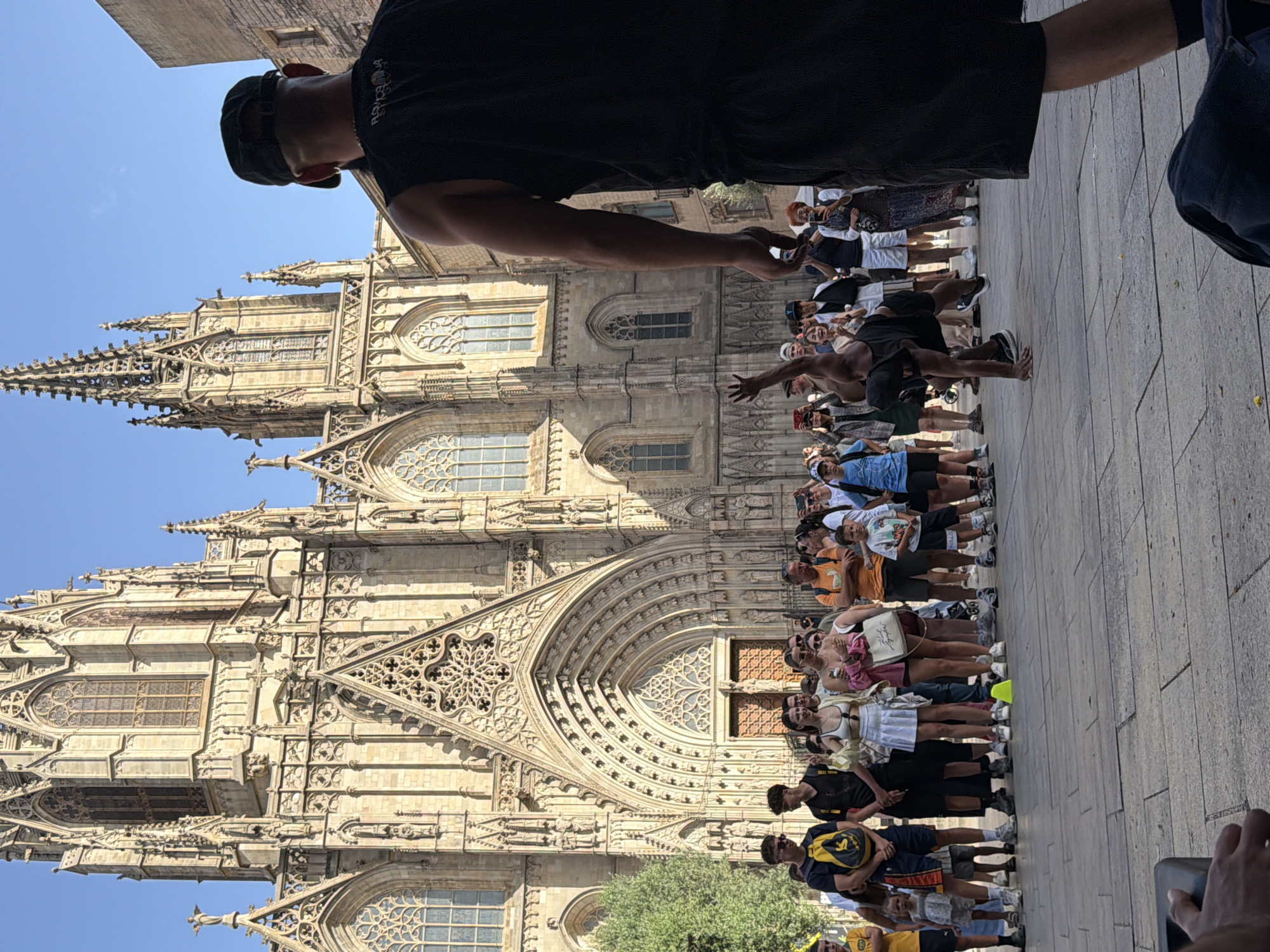 In Barcelona, Spain, in front of the Barcelona Cathedral, a breakdancer performs for a crowd of onlookers. The dancer, a Black man wearing a black sleeveless shirt and black shorts, is captured mid-move, with one hand on the ground and his body contorted. The crowd, composed of diverse individuals, watches the performance, some holding up phones to record. The Barcelona Cathedral, a magnificent Gothic structure with intricate stone carvings and tall spires, serves as a grand backdrop to the scene. The sky is clear and blue, and the ground is paved with stone tiles.