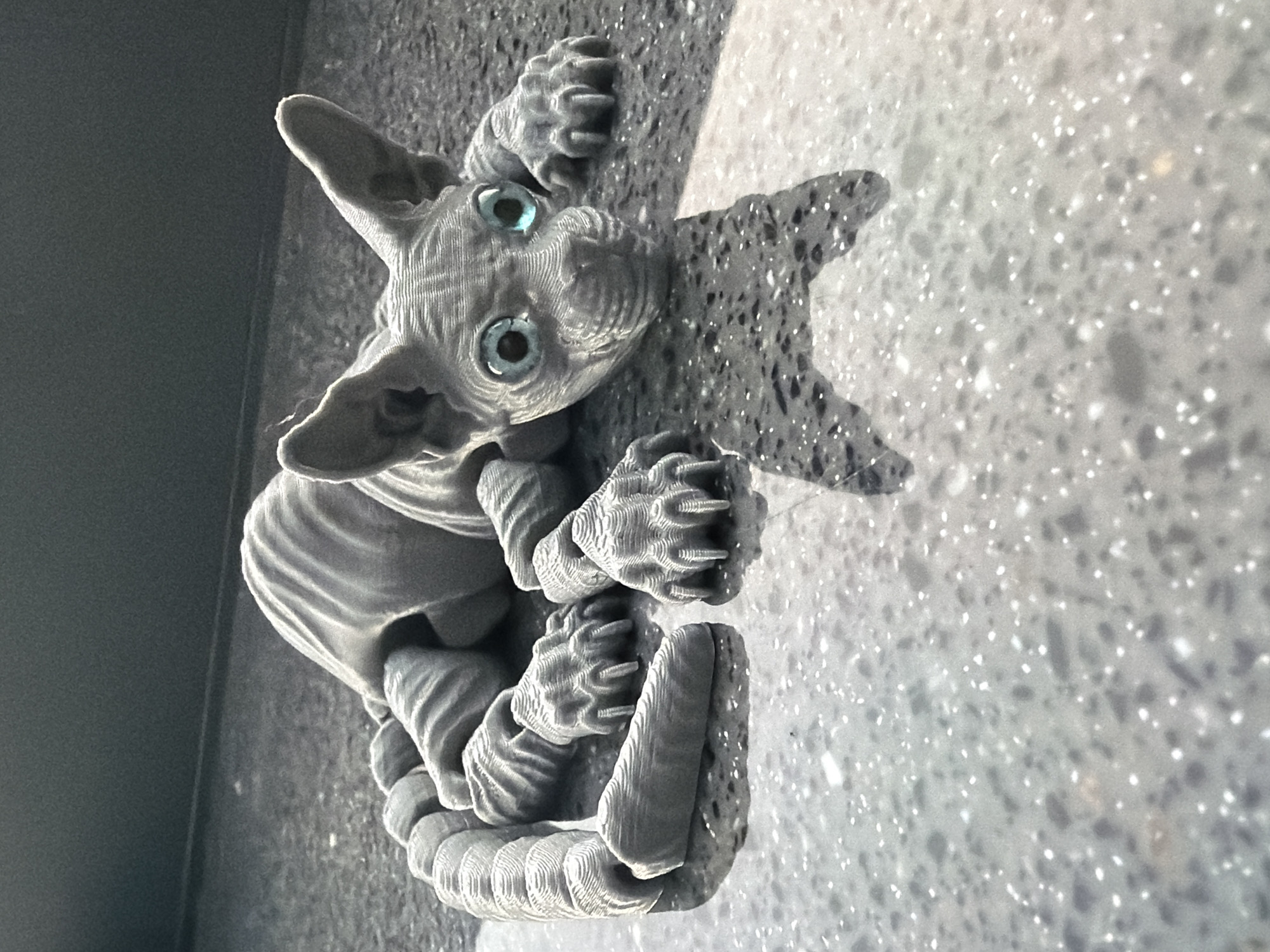 A gray, 3D-printed figurine resembling a Sphynx cat is lying on a speckled gray surface, possibly a countertop or floor, with its legs extended and its tail also stretched out behind it. The figurine has large, light blue eyes and textured details that mimic skin folds and muscle. Sunlight casts a shadow of the figurine onto the surface. The background is dark and out of focus, suggesting an indoor setting. There is no information in the image to indicate a location in Moscow, Russia.