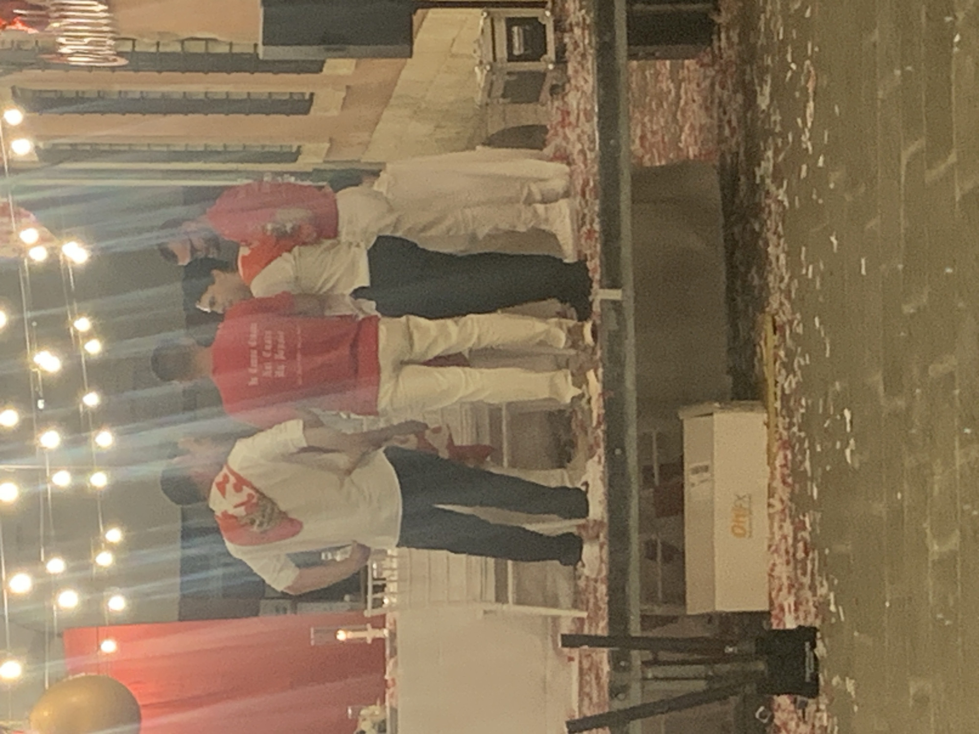 A group of five people stand on a raised platform outdoors, decorated with string lights. In the foreground, a white box with "OnFX" branding sits on the platform. Red and white confetti is scattered across the platform and the ground below. The people are dressed in casual attire, with three wearing red t-shirts and two wearing white shirts. One person in a white shirt has a red scarf draped over their shoulders. The background features a building with windows and a sign that is partially obscured. It is unclear if this image was taken in San Quirico d'Orcia, Italy.
