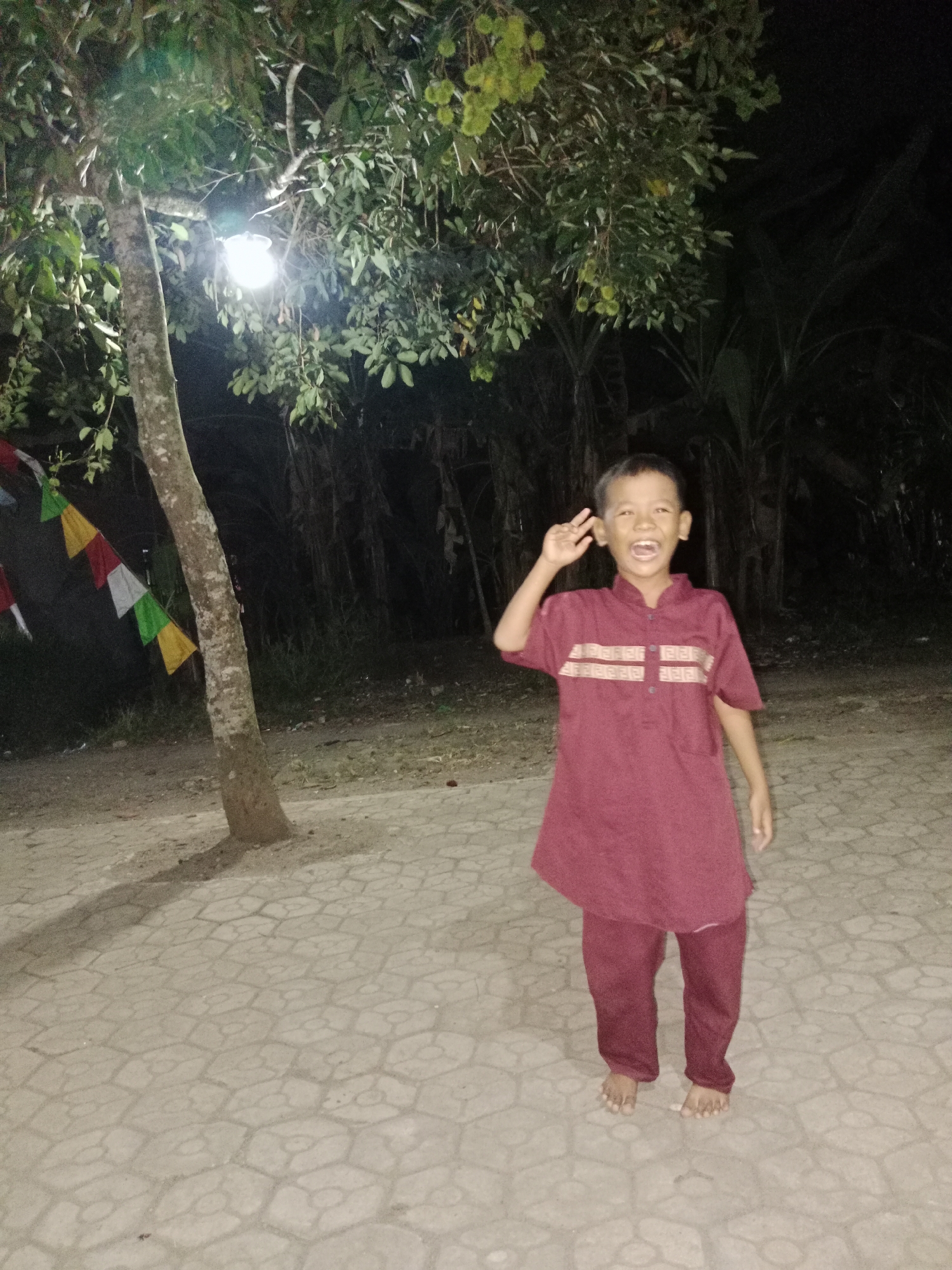A young boy is posing for a photograph at night. He is wearing a maroon tunic with decorative bands across the chest and matching maroon trousers. His left hand is raised with his index and middle fingers extended in a peace sign. He is standing barefoot on a patterned paved surface. Behind him, a tree with hanging fruits is illuminated by a light fixture. Prayer flags in various colors are strung from the tree. The background consists of dark foliage and trees. The location is identified as Parage, Indonesia.