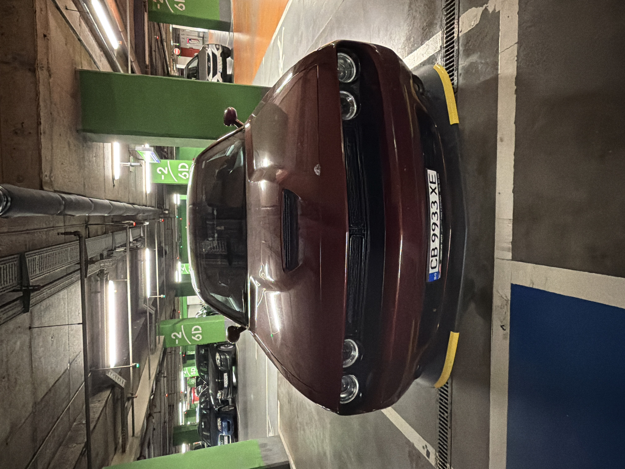 A maroon Dodge Challenger is parked in a dimly lit, underground parking garage. The car is positioned facing the camera, centered in a parking space marked with white lines. The license plate of the car reads "CB 9933 XE". The car has yellow bumper protectors on its front lower corners. In the background, other cars are visible, and green painted concrete columns are spaced throughout the garage. Overhead, fluorescent lights illuminate the concrete ceiling and exposed pipes and conduits. Signs indicating parking level and section, "-2 6D", are visible on the green columns. The overall environment suggests a typical urban parking facility.