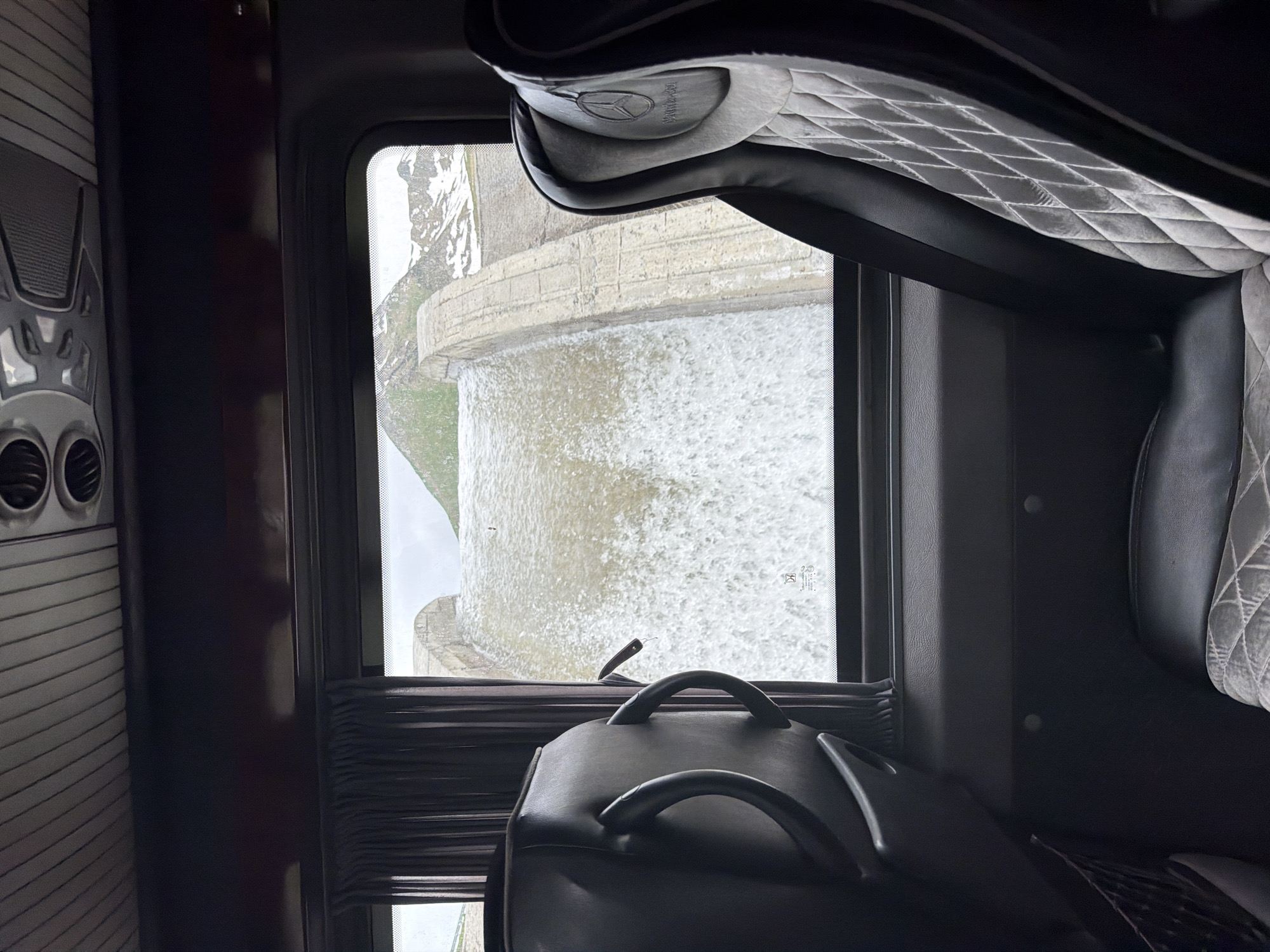 The image, taken from inside a vehicle, shows a large concrete dam with water cascading over it. The dam is curved and appears to be made of segmented concrete blocks. Mountains with patches of snow are visible in the background under a cloudy sky. In the foreground, the interior of the vehicle is visible, with a dark-colored seat on the right and the back of another seat on the left. The seat on the right has a diamond-patterned fabric on its headrest, which features a Mercedes-Benz logo. A dark curtain is drawn partially across the window on the left side. The overall scene suggests a journey through a mountainous, possibly remote, area with significant water flow.