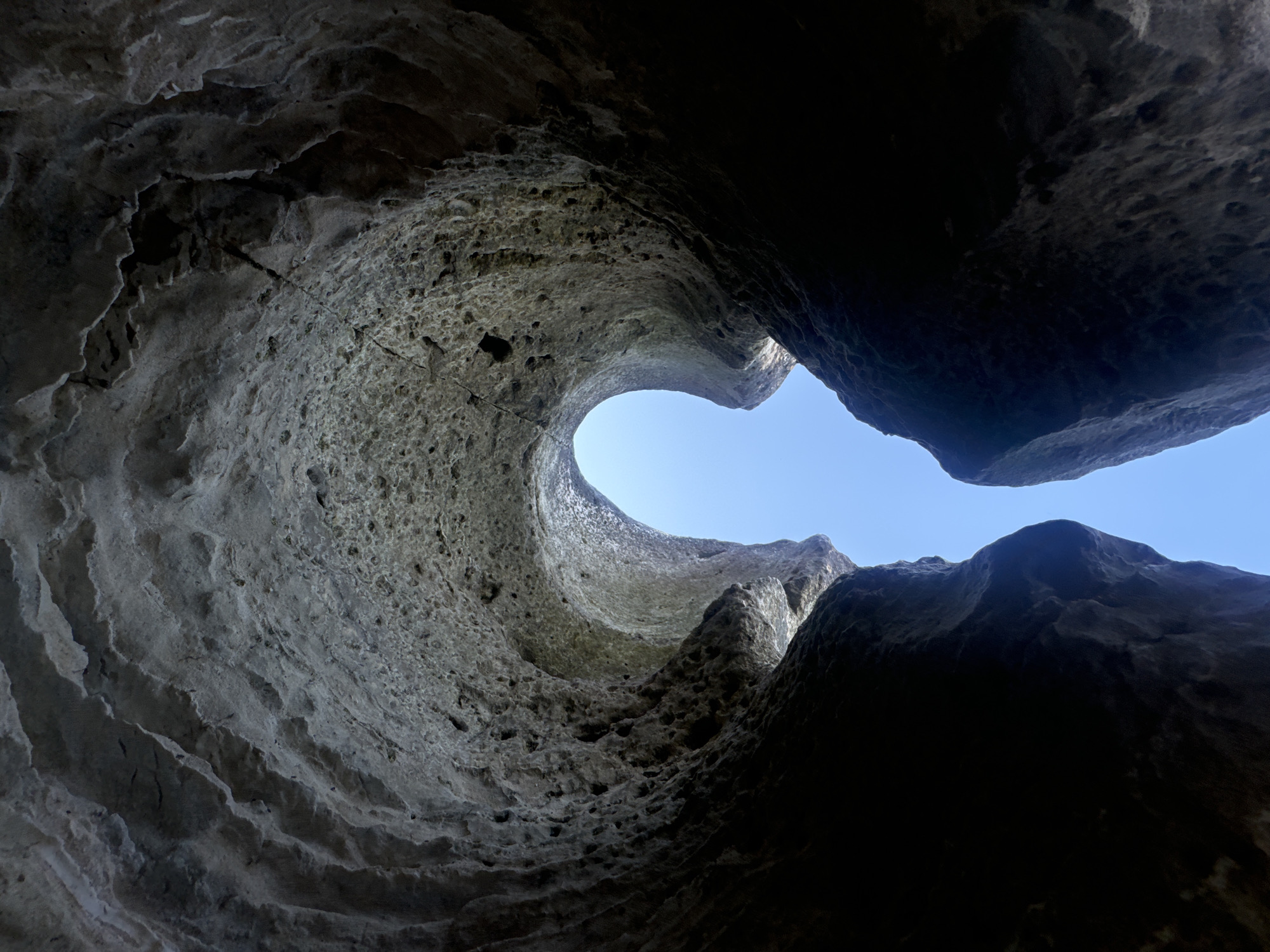 The image depicts the natural rock formations of the Bozadzhay mountain-valley, located near the city of Aktau, Kazakhstan. The viewpoint is from within a natural hollow or cave-like opening in the rock, looking upwards towards the sky. The rock surfaces are rough and textured, with visible layers and pockmarks, suggesting erosion over time. The opening is framed by dark, shadowy rock on either side, creating a distinct U-shape with a bright blue sky visible through the aperture. The lighting suggests it is daytime, with the sky being a clear, cloudless blue. There are no people or man-made objects visible in the frame, focusing entirely on the geological formations.