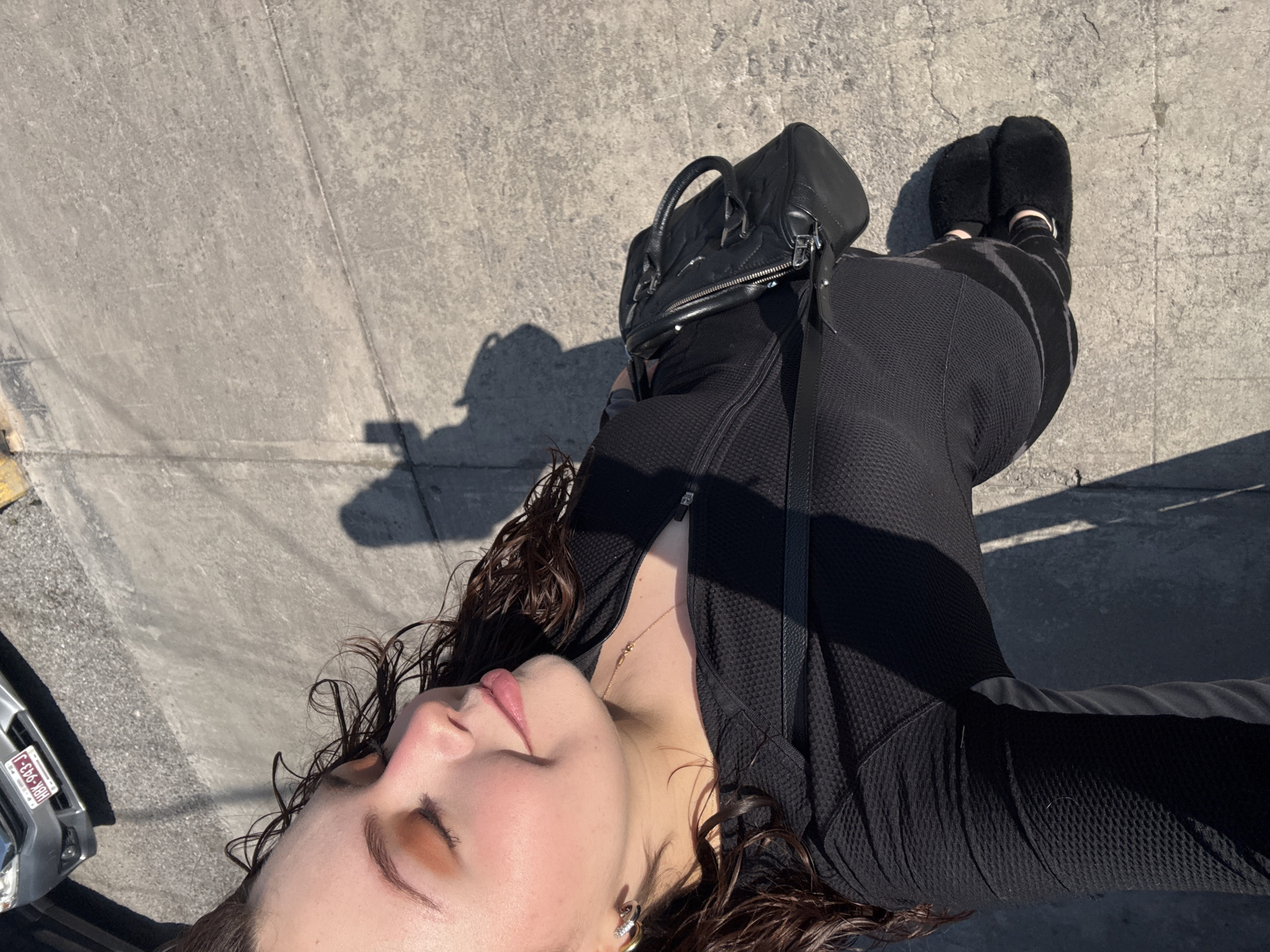 A woman is standing on a city street in Puebla, Mexico, with a black bag hanging from her shoulder. She is wearing a black jacket, pants, and furry black slip-on shoes. Her hair is curly and damp, and she is looking upwards with her eyes closed. The street is made of concrete, and there is a car parked nearby. The license plate on the car is HBM-933-J.