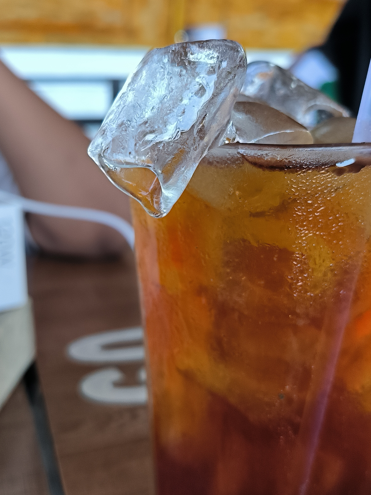 A close-up shot shows a glass of iced tea. The glass is sweating with condensation. Several ice cubes fill the glass and one large cube is perched precariously on the rim, partially hanging over the edge. A white drinking straw is visible within the glass.
In the background, out of focus, is part of a table with some writing on it, a glimpse of a person's arm, and a small portion of the surroundings, possibly another table or chair.
Given the location of Matang, Indonesia, the setting appears to be an outdoor cafe or restaurant setting.