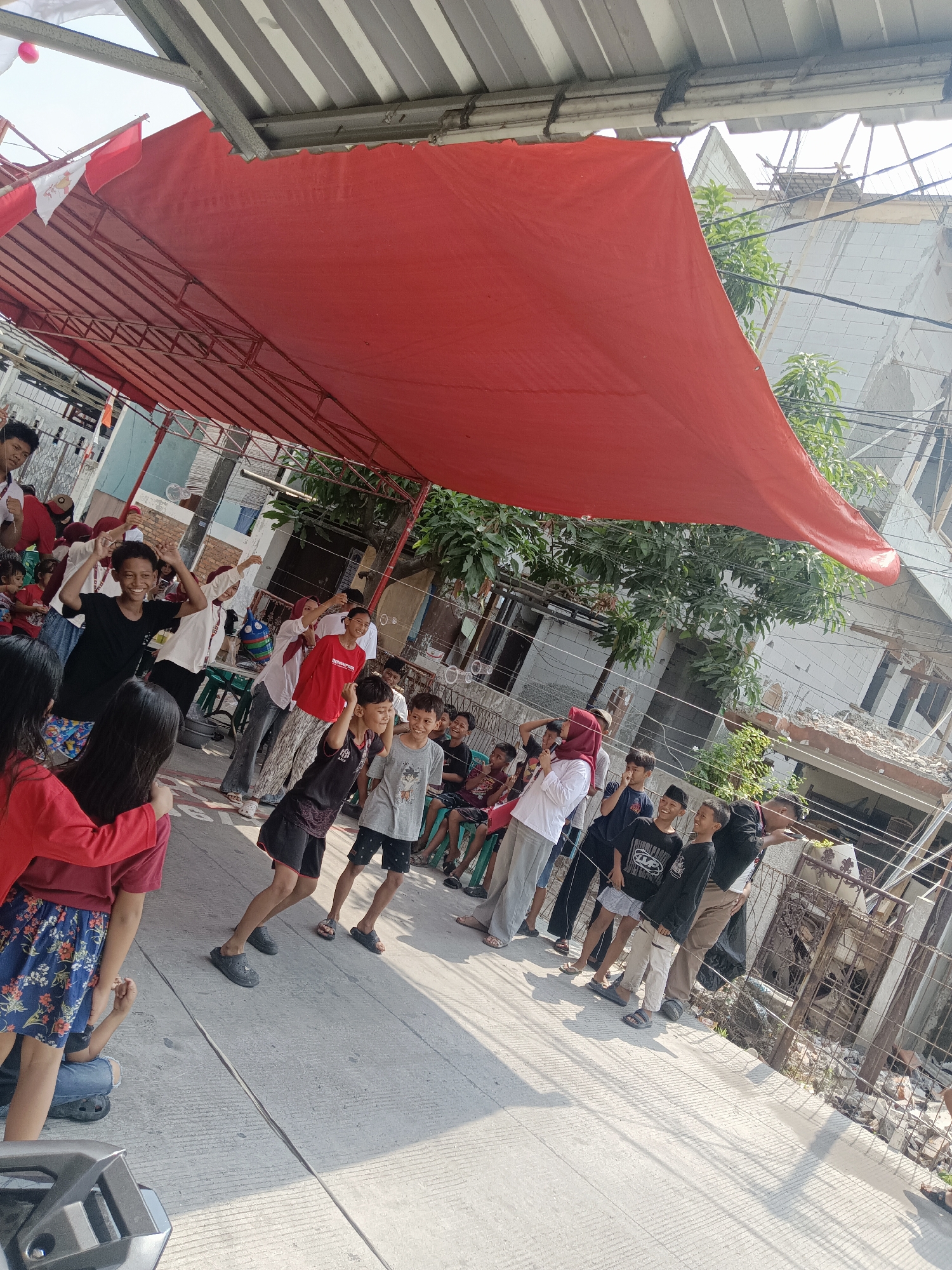 In North Jakarta, Indonesia, children are participating in an outdoor event under a large red tarp canopy. The scene appears to be taking place on a street or open paved area, possibly in a residential neighborhood given the presence of a partially damaged building in the background. The children, dressed in casual clothing, are engaged in various activities, with some actively playing and others observing. One group of boys in the foreground are smiling and appear to be posing for the camera. Several people, including adults and children, are gathered under the canopy, suggesting a community or festive gathering. The environment shows signs of development or repair, with exposed bricks and scaffolding visible on the damaged building. The overall atmosphere seems lively and communal, possibly during a local celebration or event.