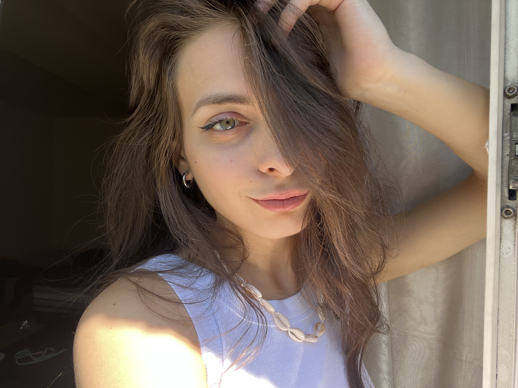 A young woman with long, brown hair and green eyes poses for a selfie. She is wearing a white tank top and a seashell necklace. Her left hand is in her hair, and her right arm is bent at the elbow, resting on her head. The woman is looking at the camera with a slight smile. The background is dark, with sunlight streaming in from the left, illuminating her face and shoulder. The image is taken indoors, likely in a room with a window. Krasnoyarsk, Russia.
