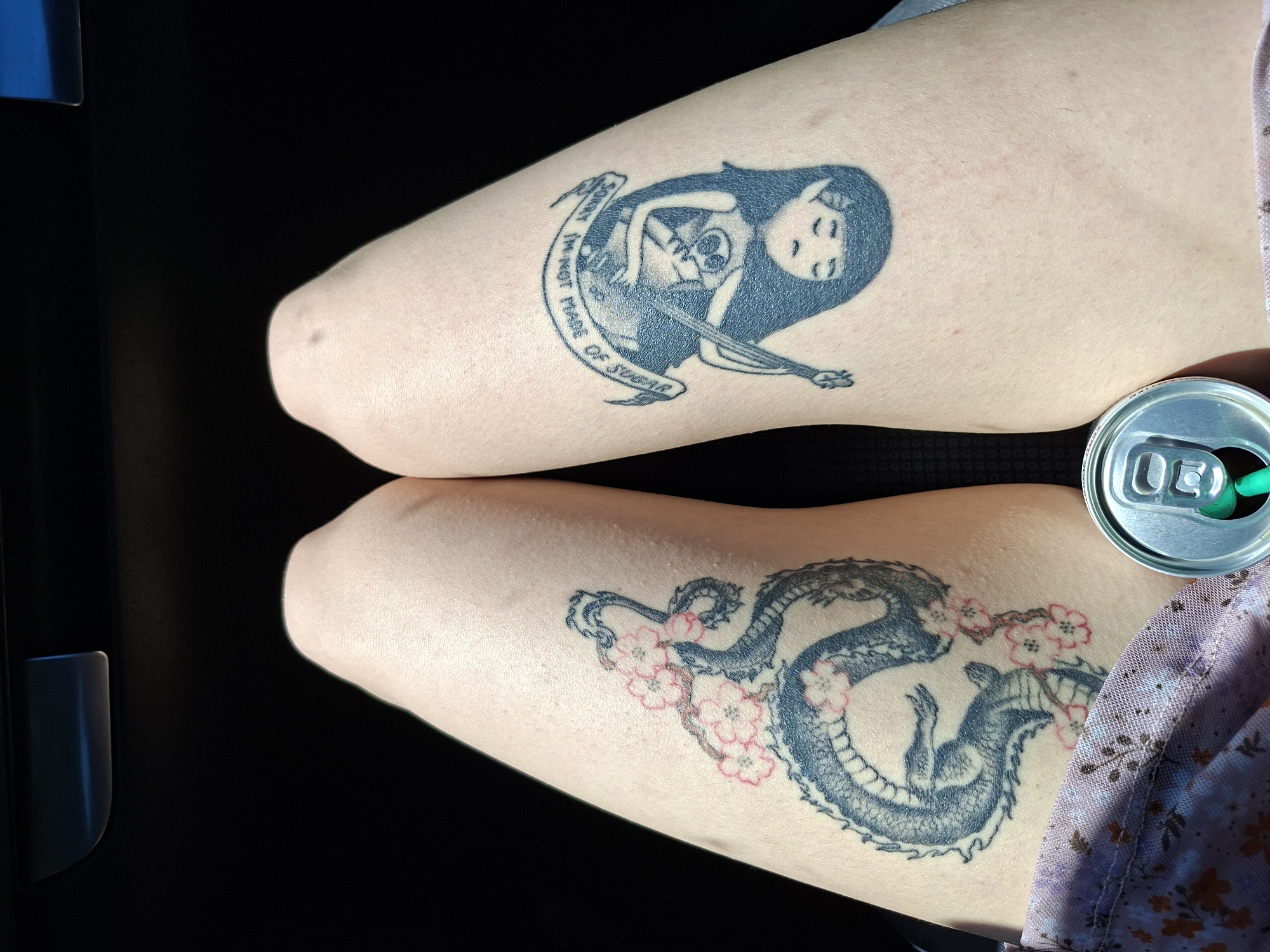 The image shows two human legs with tattoos. On the left leg is a tattoo of a female figure cradling a skeleton while playing a stringed instrument. A banner surrounds the image with the text "Sorry I'm not made of sugar". On the right leg, there is a tattoo of a dragon intertwined with cherry blossom branches. A partially visible can of a drink with a green straw rests next to the right leg. The background is dark, suggesting an indoor setting, possibly a vehicle. The image does not provide any information regarding the location in the city of Kamburovo, Bulgaria.