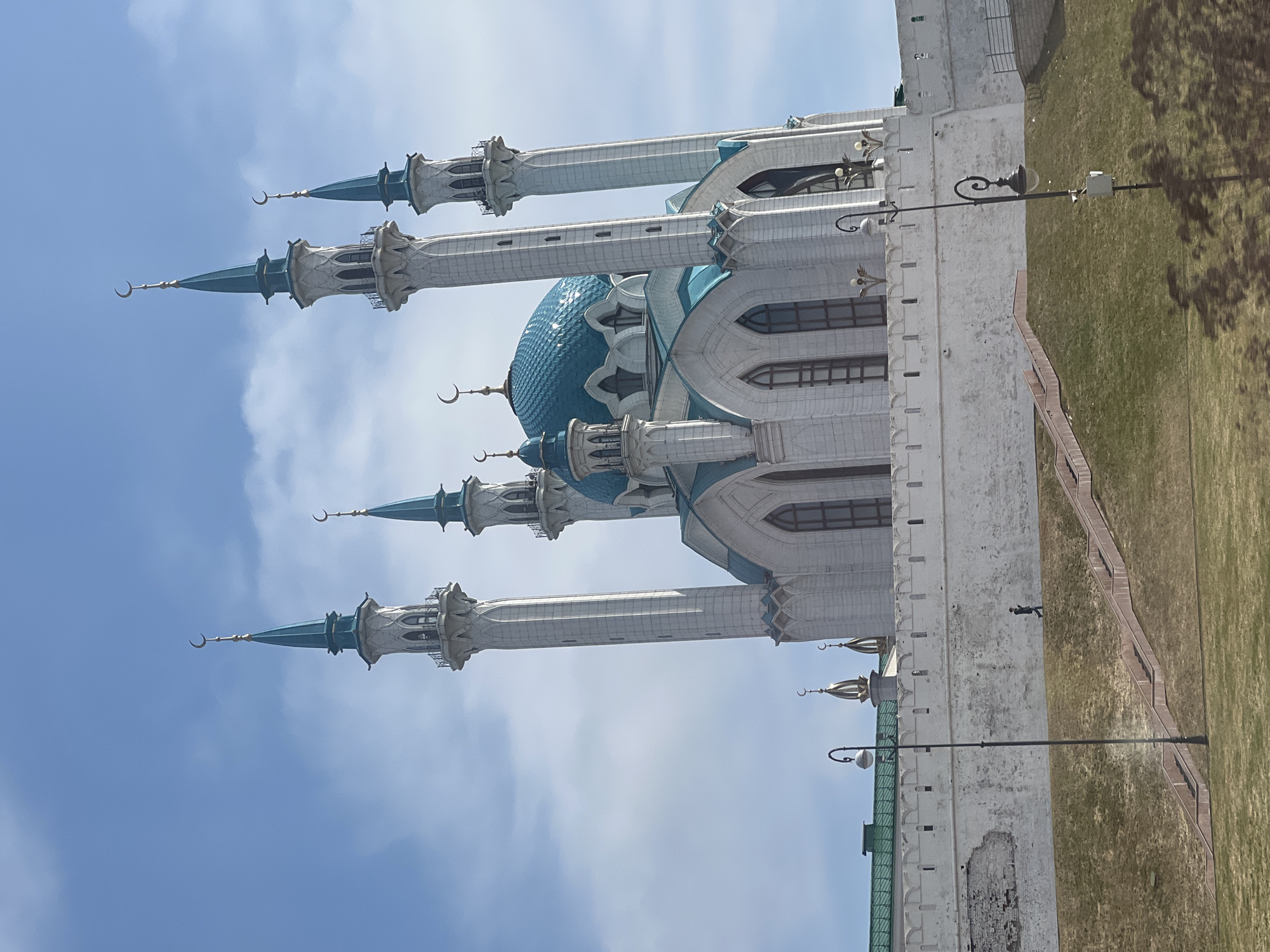 The image shows the Kul Sharif Mosque in Kazan, Russia. The mosque is a large, white building with blue domes and minarets. The image is taken from a low angle, looking up at the mosque. The sky is blue with some clouds. The foreground shows a grassy area with a path. A white wall is visible, along with antique styled street lamps.