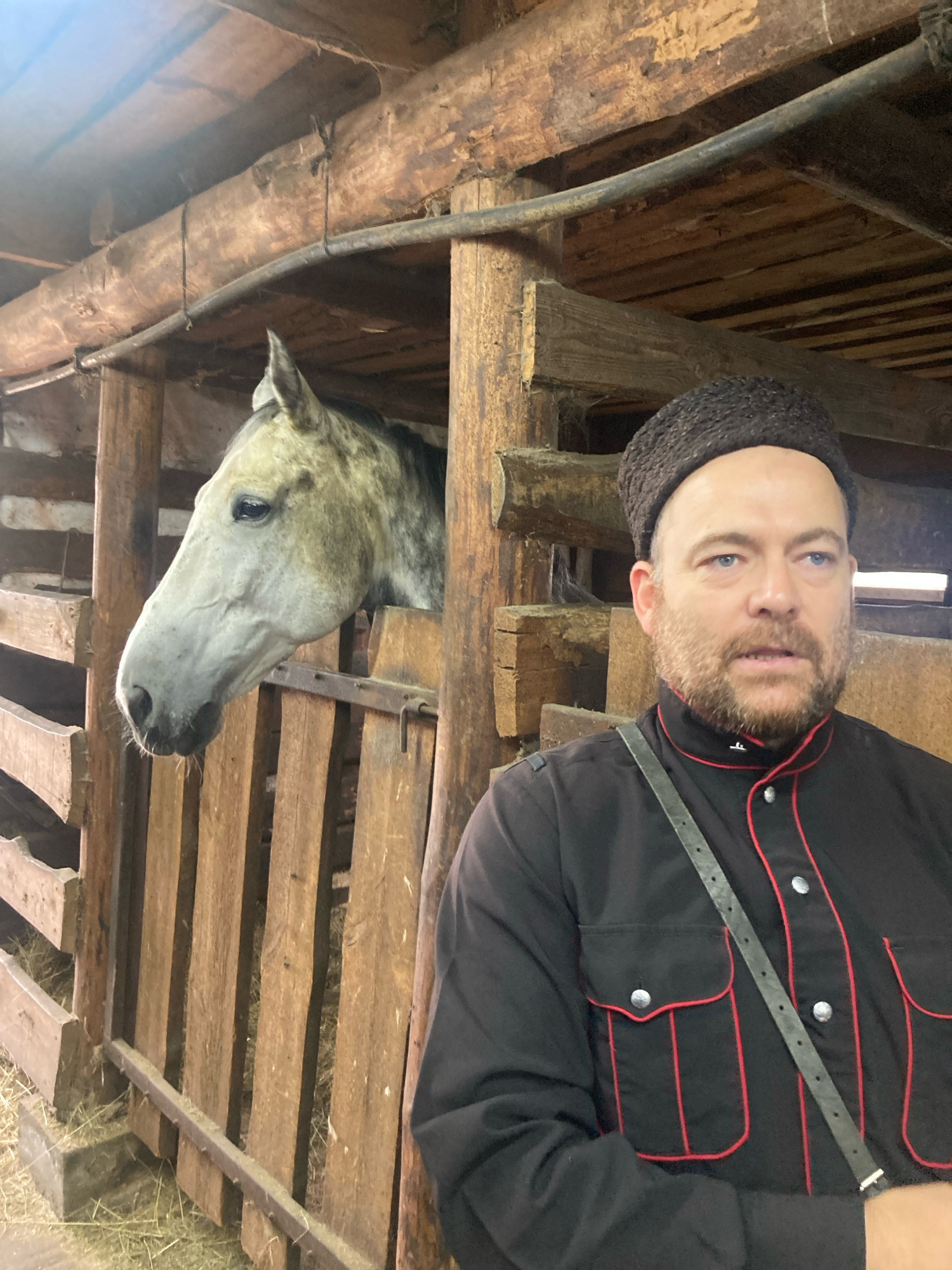 A medium shot captures a man and a horse in a rustic wooden stable. The man, positioned on the right side of the frame, wears a black, button-down shirt with red trim and a dark, textured cap. He has a beard and blue eyes and appears to be looking off to the right. A dark strap crosses his chest. To the left of the man, a dappled grey horse's head and neck are visible, peering out from a stall within the stable. The stable is constructed of rough-hewn wooden beams and planks, with hay scattered on the floor. The lighting suggests an indoor setting, possibly during the day. The background reveals more of the stable's wooden structure, including overhead beams and a wooden fence. The scene is devoid of any explicit indication of the city of Тэльмінскі сельскі Савет, Belarus.