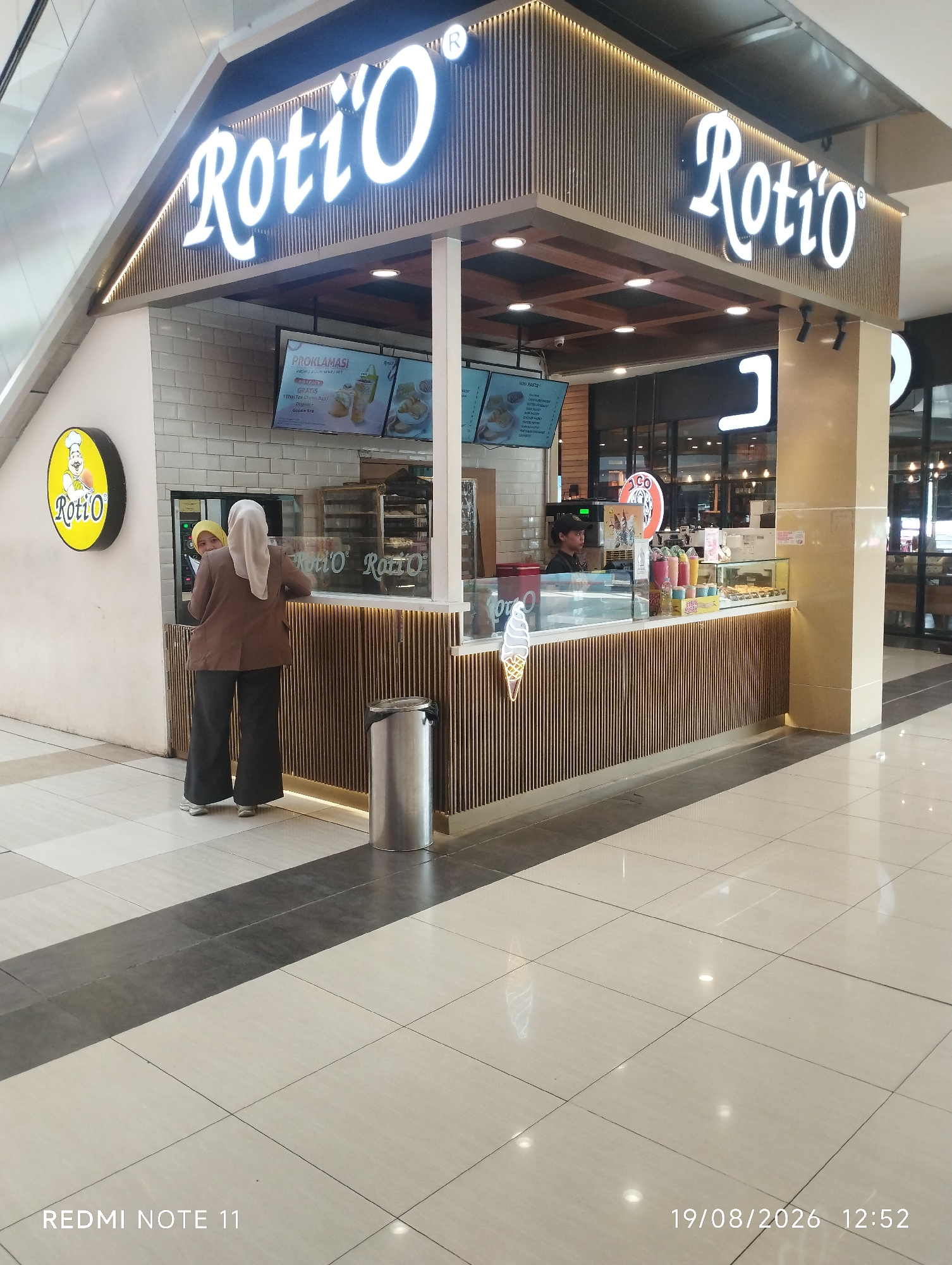 In Karawang, Indonesia, a Roti'O store is depicted in a shopping mall. The store features a prominent Roti'O logo and a display counter with various food items. Two individuals are seen at the counter, with one ordering while the other appears to be an employee. The mall environment is evident with tiled flooring and overhead lighting. The image captures a commercial scene, highlighting the presence of a popular food franchise in the city.
