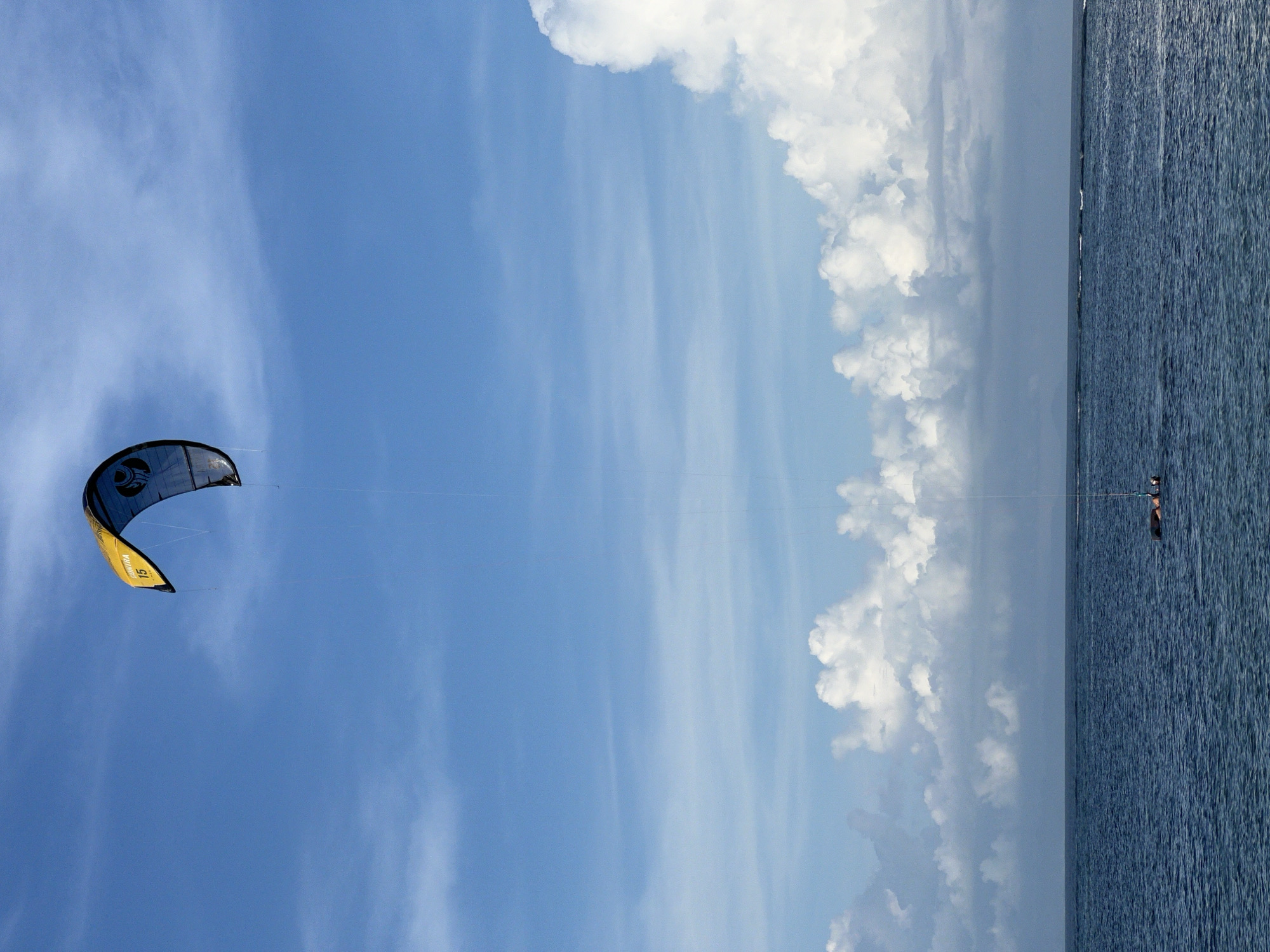 A kitesurfer is visible in the water, attached to a black and yellow kite in the blue sky. The sky also features large white clouds. The scene is likely located in Denpasar, Indonesia, given the information provided. The water's surface appears slightly choppy.