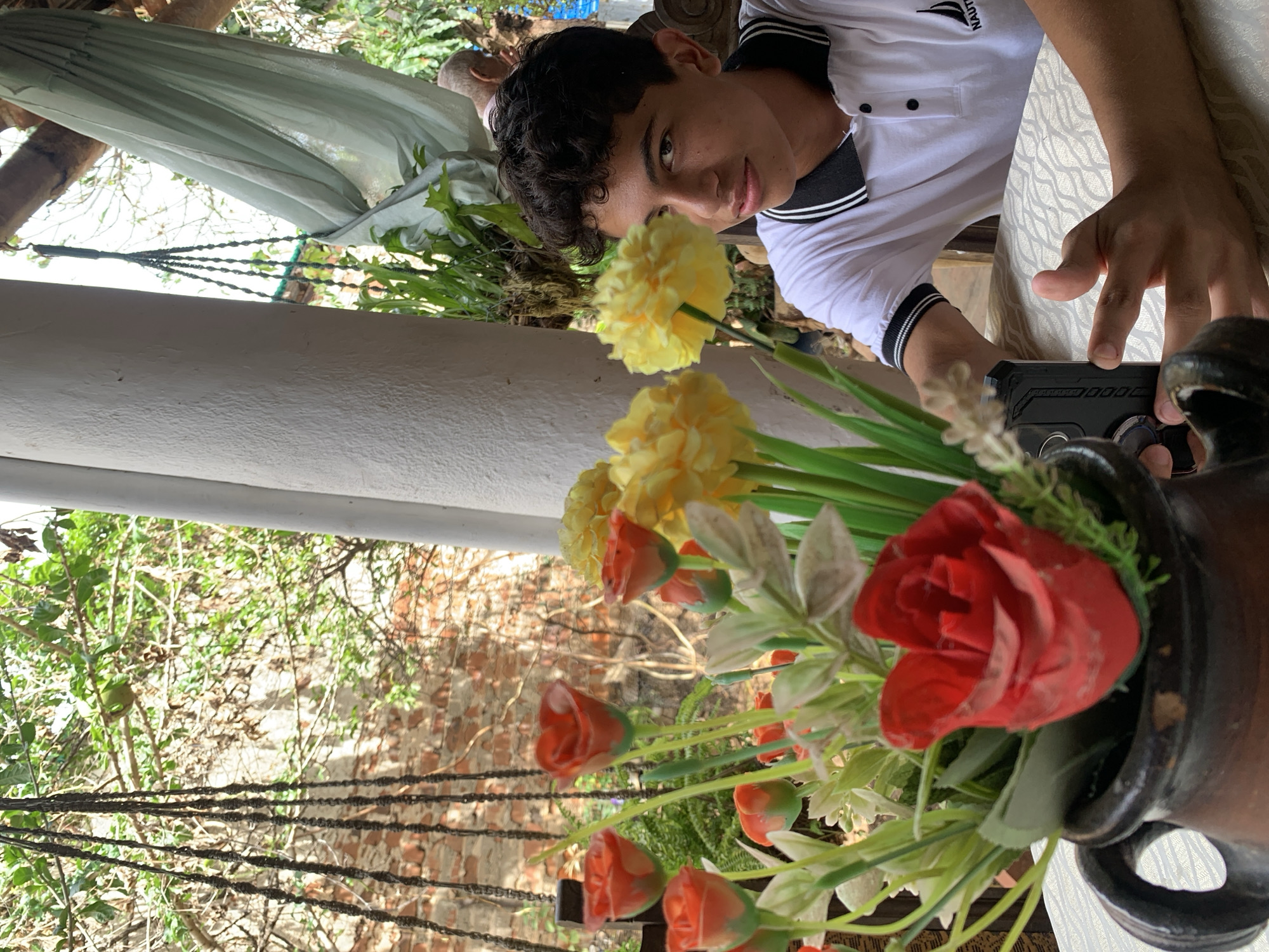 The image depicts a young male seated at a table in a semi-outdoor setting in Tacarigua, Venezuela. The foreground features a dark brown ceramic pot containing a bouquet of artificial flowers, primarily red roses (some in bud, one fully open) and yellow pom-pom-like blooms, accompanied by green foliage and light-colored floral elements. The table is covered with a white tablecloth featuring a wavy, geometric pattern.
The main subject, a young male with dark, curly hair and light brown skin, is positioned slightly right of center, looking directly at the viewer with a neutral expression. He wears a white polo shirt with a black collar and black striped armbands, featuring a "NAUTICA" logo on the right chest. His right hand holds a black smartphone encased in a rugged protective cover. His left forearm and part of his upper body are also visible.
The background consists of a vibrant outdoor environment. To the left, a portion of a rustic brick wall is visible, partially obscured by lush green foliage and hanging dark braided ropes, possibly from a planter. A thick white support pillar is prominent on the left. To the right of the main subject, dense green vegetation and overhead wooden beams are visible. A light green, folded fabric, possibly a hammock or curtain, hangs from an overhead beam in the upper right. A blurry, older male figure with short gray hair is partially discernible in the distant background on the right. The scene is brightly lit by natural light.