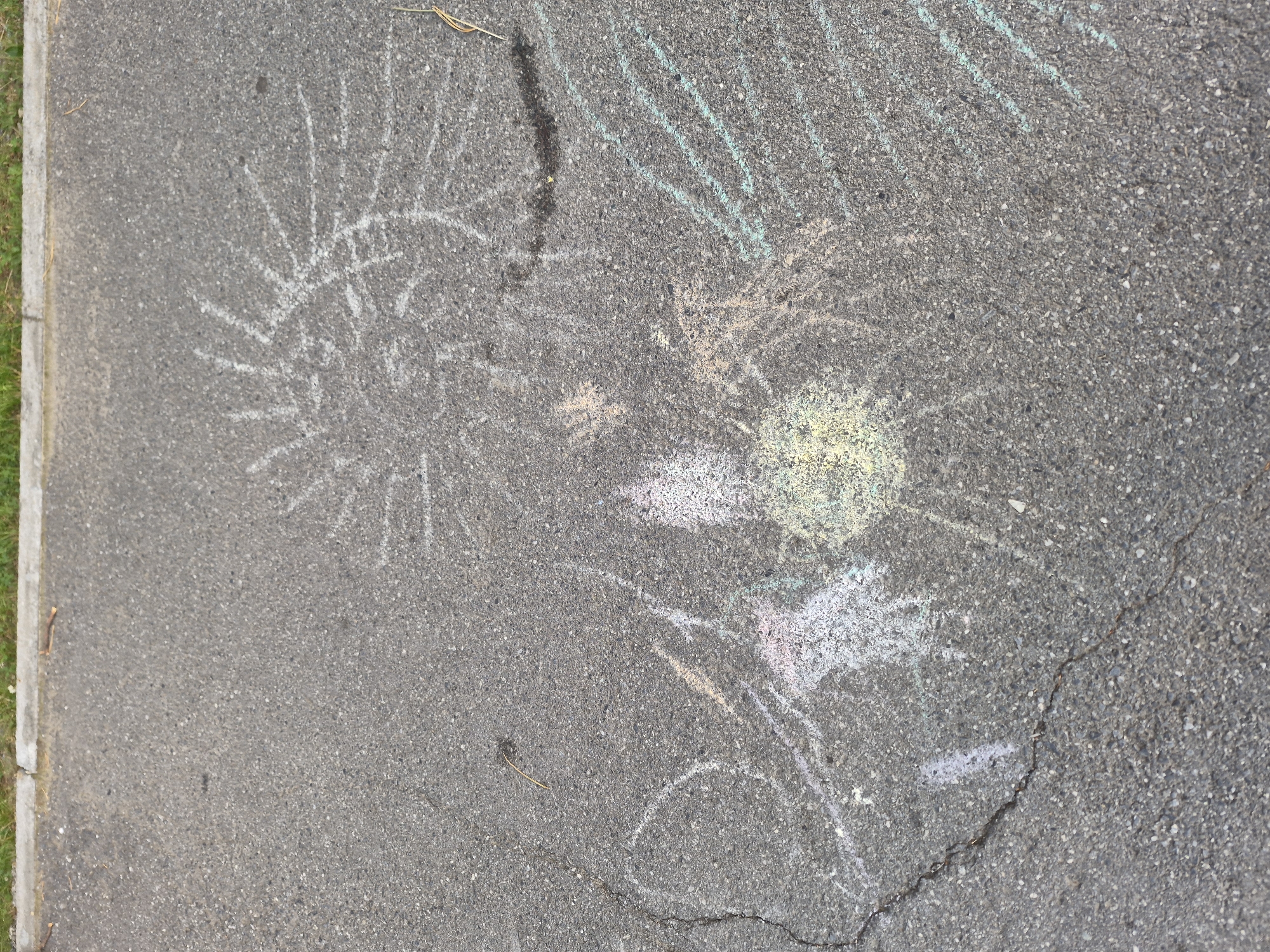 The image shows chalk drawings on asphalt. The drawings appear to be abstract and colorful, with one resembling a sun or a starburst in white chalk. Other chalk marks include streaks of green, yellow, pink, and white, suggesting a child's artwork. The asphalt is grey and appears to be part of a sidewalk or driveway, with a concrete curb and grass visible on the left edge. A few dry twigs or pieces of straw are scattered on the ground. The lighting suggests it is daytime. There is no visible text in the image.