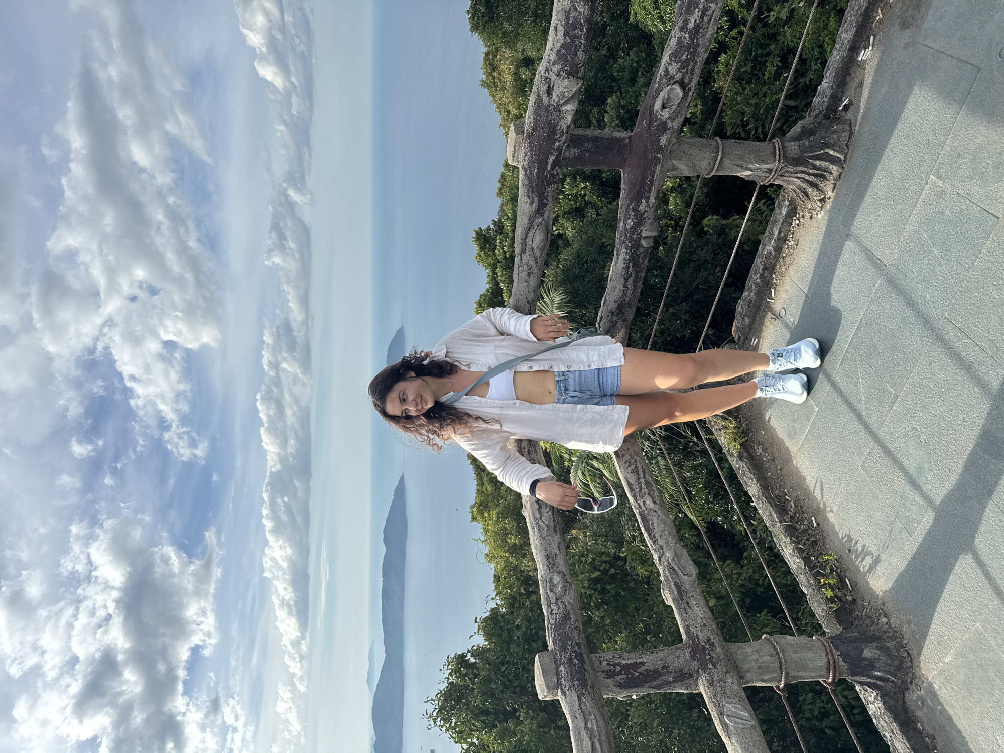 A woman stands on a raised, paved viewing platform overlooking a bay with islands in the distance. The platform is enclosed by a rustic wooden railing made of rough-hewn logs and concrete posts, with rope detailing. The woman, wearing a white crop top, denim shorts, an unbuttoned white linen shirt, and light blue sneakers, leans on the railing with one hand. She holds a pair of white-rimmed sunglasses in her other hand. A light blue crossbody bag is slung over her shoulder. Behind her, lush green foliage descends the slope towards the water. The sky is filled with dramatic, scattered clouds against a bright blue expanse. This scene appears to be at the Son Tra Peninsula in Da Nang, Vietnam, likely at one of the viewpoints offering panoramic vistas of the coastline and the East Sea.