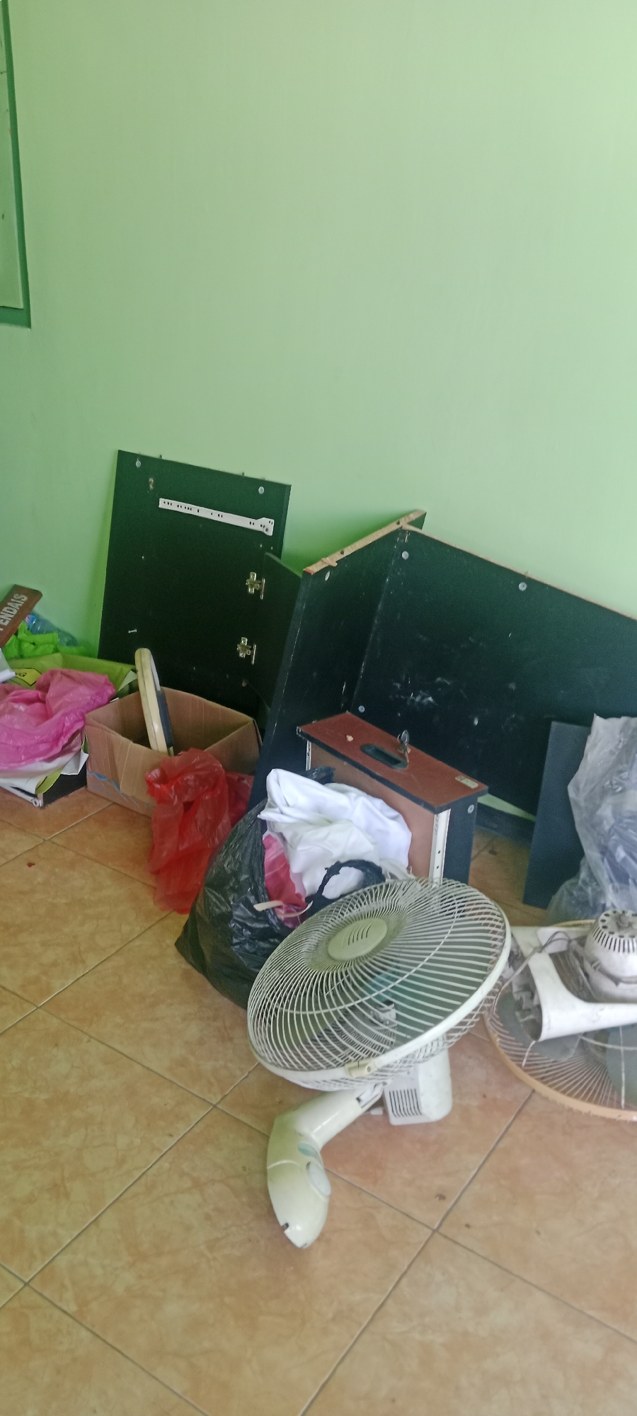 This image depicts a cluttered room, likely a storage or unused space, with the overall impression of disarray. The walls are painted a pale, light green color. The floor is tiled with square, light brown tiles.
In the foreground and mid-ground, there are several objects contributing to the clutter. A white oscillating fan with a white base and a metal safety grill is prominently placed on the floor, angled towards the right side of the frame. To the right of this fan, partially visible, is another fan with a similar design, suggesting multiple fans are present.
Several pieces of what appear to be disassembled or discarded furniture are leaning against the wall. These are dark-colored, possibly black or very dark wood, with visible screws and hinges. One piece has a white metal drawer slide attached. Another piece is a small, dark brown cabinet with a red-brown top and a handle.
Surrounding the furniture and fans is a collection of miscellaneous items. There is a large black trash bag filled with various items, including white and pink fabrics, and possibly other discarded materials. Next to it, a red plastic bag is visible, as well as a cardboard box with a light-colored rim. Pink and white plastic bags or fabric are also strewn about to the left. A white circular object, possibly a part of something, is also visible.
The overall scene suggests a room that is not currently in use or is undergoing a clean-out or renovation. The lighting is moderate, casting some shadows but not obscuring the details of the objects. The image does not provide any specific indicators to pinpoint its location within the city of Tuban, Indonesia, beyond being an indoor scene.