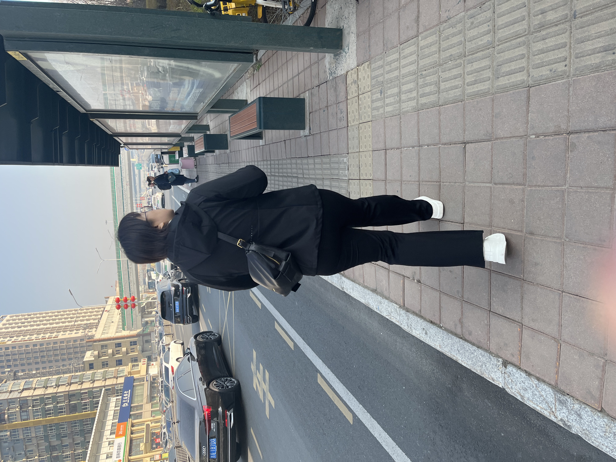 This image, captured in Changchun City, China, presents an urban street scene from the perspective of someone standing on a sidewalk. The primary subject is a person, seen from behind, wearing a black jacket, black pants, and white shoes, and carrying a black shoulder bag. They are standing on a paved sidewalk, which includes sections of tactile paving for the visually impaired, looking towards a street filled with traffic.
To the left, a bus stop shelter runs along the sidewalk, equipped with benches and transparent panels, possibly for advertisements. Further down the sidewalk, other individuals are visible, some with luggage, suggesting a busy public transport area.
The street is active with vehicles, including several cars. A prominent black Audi sedan displays the license plate "吉A B170U", and another car in the background has "吉A 87Y02". Traffic lights showing red signals are visible. White road markings and Chinese characters spelling "靠站" (Kàozhàn), meaning "stop/pull over (at a station)", are painted on the asphalt.
In the background, a cityscape unfolds with numerous multi-story buildings under a clear, bright sky, indicating a daytime setting. One building features a blue sign with the Chinese characters "吉林银行" (Jilin Bank). The scene depicts a vibrant city environment with daily commuting and urban activity.