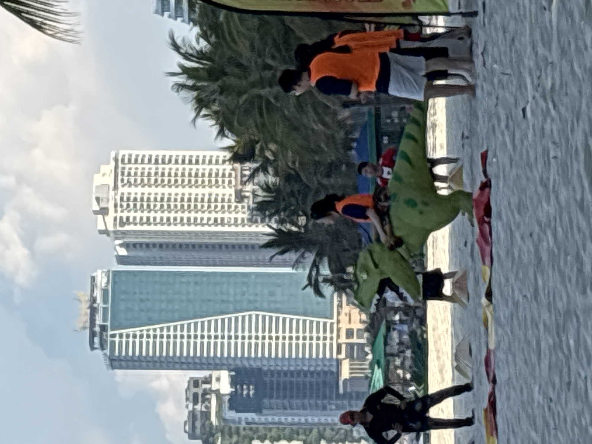 This media file captures a lively scene on a sandy beach in Đà Nẵng, Vietnam. The image is oriented vertically, revealing a group of people engaged in an activity, possibly a competition or a themed event. Several individuals are wearing bright orange vests, suggesting they are participants or organizers. One person is dressed in a green dinosaur costume, adding a playful and whimsical element to the gathering.
In the background, the urban landscape of Đà Nẵng is visible, with modern high-rise buildings contrasting with the natural beach setting. Palm trees line the beach, indicative of a tropical environment. The sky appears to be clear with scattered clouds, suggesting fair weather. The lighting suggests it is daytime, likely late morning or early afternoon, given the bright sunlight casting shadows.
The people appear to be interacting with each other, with some focused on the dinosaur costume and others milling about. The overall mood seems energetic and positive, with a sense of community and shared enjoyment. The presence of what looks like event signage or banners in the background further supports the idea that a specific event is taking place. The sandy texture of the beach and the calm water in the distance are characteristic of a coastal setting. No visible text can be deciphered in the provided image.