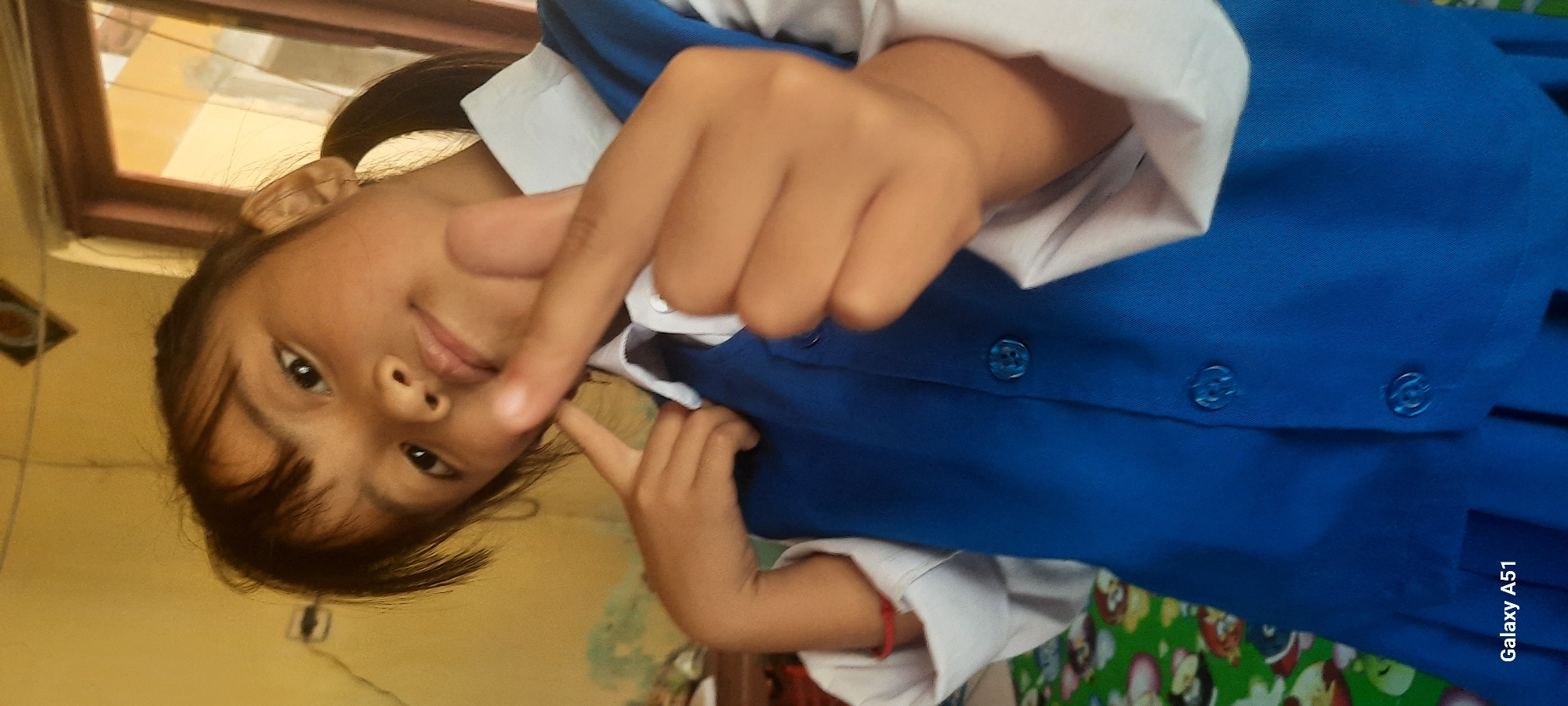 In Ploso, Indonesia, a young girl, likely of Indonesian ethnicity, poses for a photograph in what appears to be a classroom or home setting. She is wearing a school uniform, consisting of a white collared shirt and a blue vest with blue buttons. Her hair is dark and styled in pigtails. She is looking towards the camera with a slight smile and her right hand is positioned near her face, making a heart shape with her thumb and index finger. Her left hand is touching her cheek. The background is predominantly yellow, with a hint of green and brown to the left and right respectively, possibly indicating walls or furniture. A patterned fabric, possibly a tablecloth or bedspread, is visible at the bottom left. The image has a "Galaxy A51" watermark in the bottom left corner.