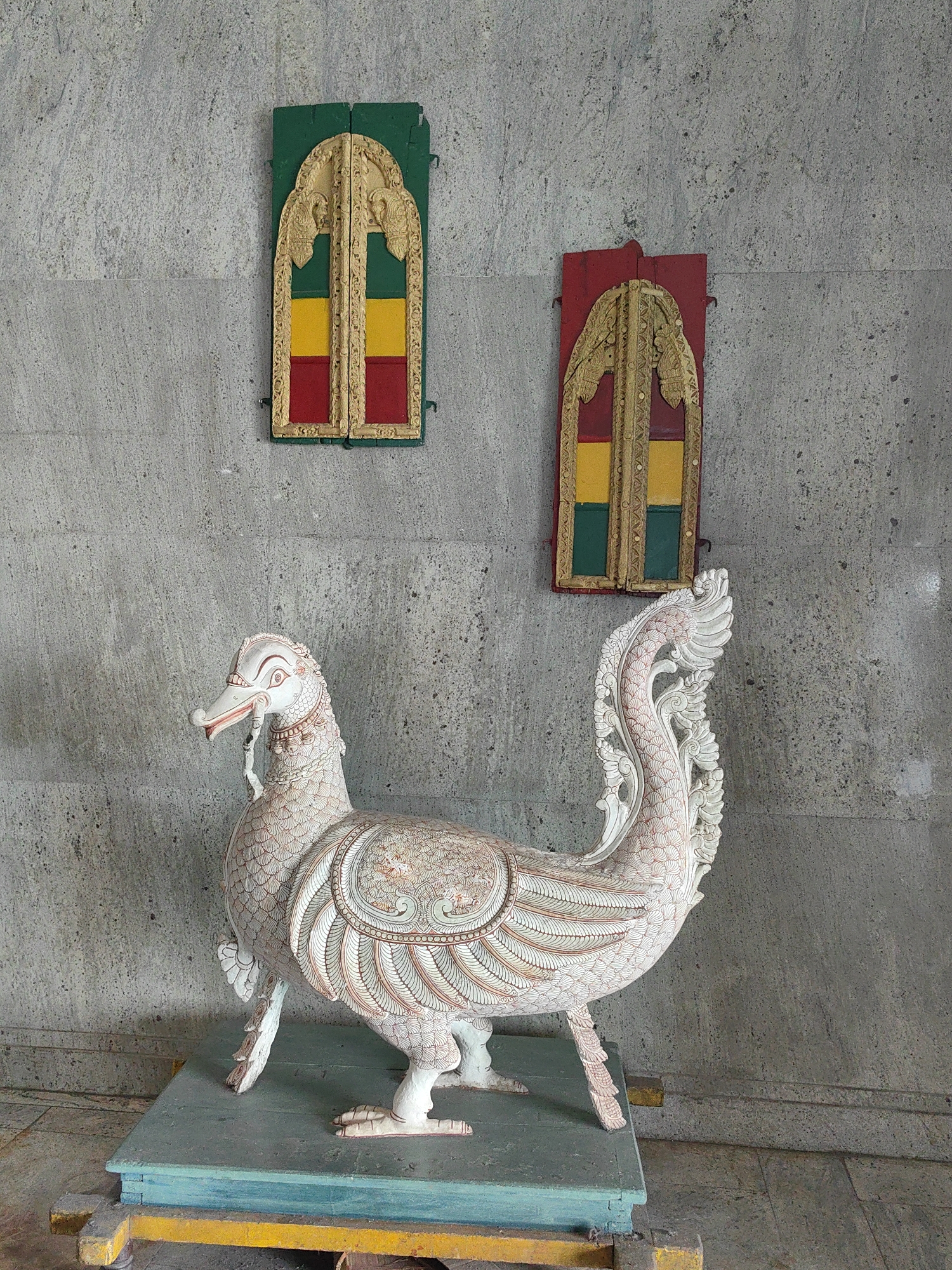 The image shows a carved wooden statue of a mythical bird, possibly a swan or peacock, standing on a platform. The statue is intricate, with detailed carvings depicting feathers and ornate patterns. The bird is predominantly white with delicate red and light green accents. The platform it stands on is a layered structure with colors of green, blue, and yellow.
Behind the statue, mounted on a textured gray wall, are two ornate, rectangular decorative panels. Both panels feature a similar arched design at the top, with intricate golden carvings. The panels are divided into sections filled with bright colors: one panel has green at the top, followed by yellow, and then red at the bottom. The other panel has red at the top, followed by yellow, and then green at the bottom.
The overall scene appears to be indoors or in a covered outdoor area, given the presence of the wall and the controlled lighting. The style suggests a cultural or religious setting, potentially a temple or a museum exhibit. Without additional information or context, pinpointing the exact location within Mumbai is not possible based solely on the visual elements.