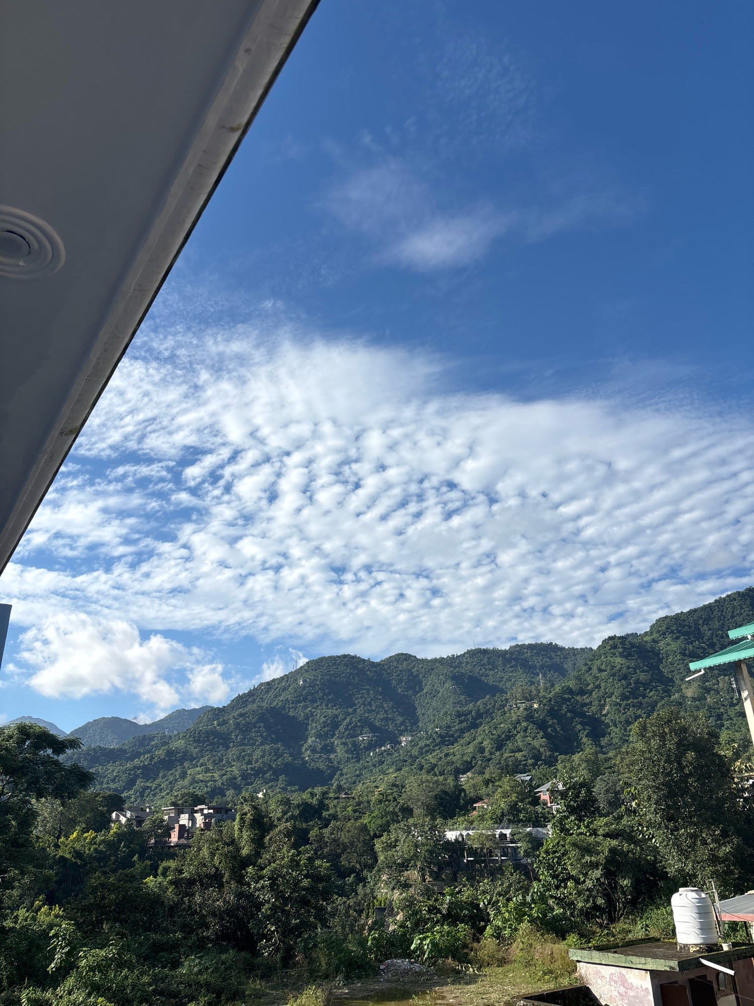The image captures a serene mountain landscape in Bhatta, India, under a bright blue sky dotted with scattered white clouds. A prominent dark overhang, likely from a building's roof, occupies the top left portion of the frame. Below this, the lush green slopes of rolling mountains dominate the scene, covered in dense trees and vegetation. Several buildings are nestled among the hills, appearing as clusters of houses and structures partially obscured by the foliage. On the right side of the frame, a white water tank is visible on top of a building, along with a portion of a green-roofed structure. The foreground shows a patch of overgrown grass and some debris, suggesting a natural, somewhat uncultivated area. The overall atmosphere is peaceful and scenic, highlighting the natural beauty of the mountainous region.