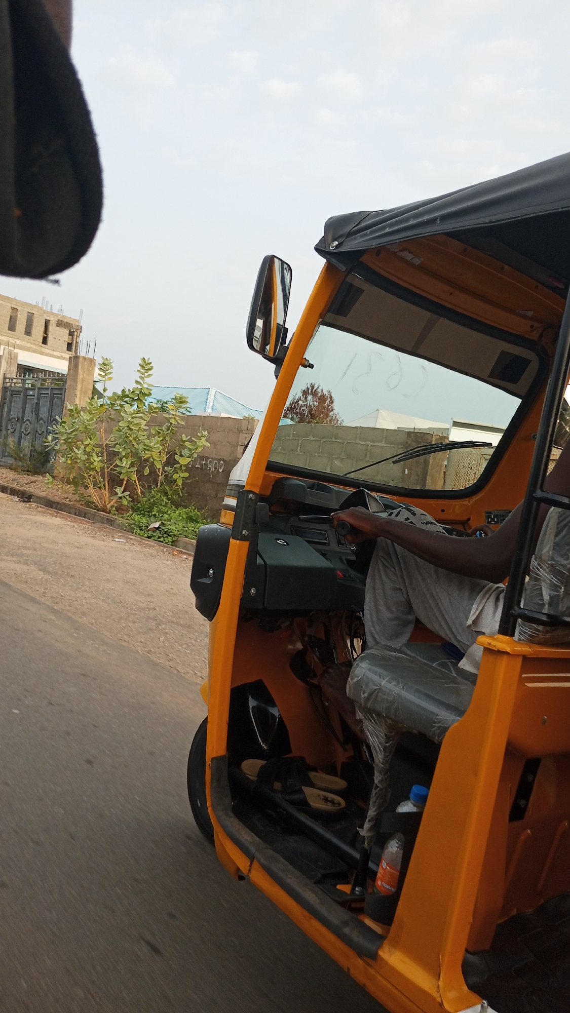 A partially visible, dark-skinned person is driving a bright yellow, three-wheeled taxi on a paved road in Adankolo, Nigeria. The vehicle has a black canopy top and a side mirror. A water bottle is visible inside the taxi, and a pair of sandals rests on the floor. In the background, there is a building with a blue roof. A gray brick wall and a gate are also visible. The sky is overcast.