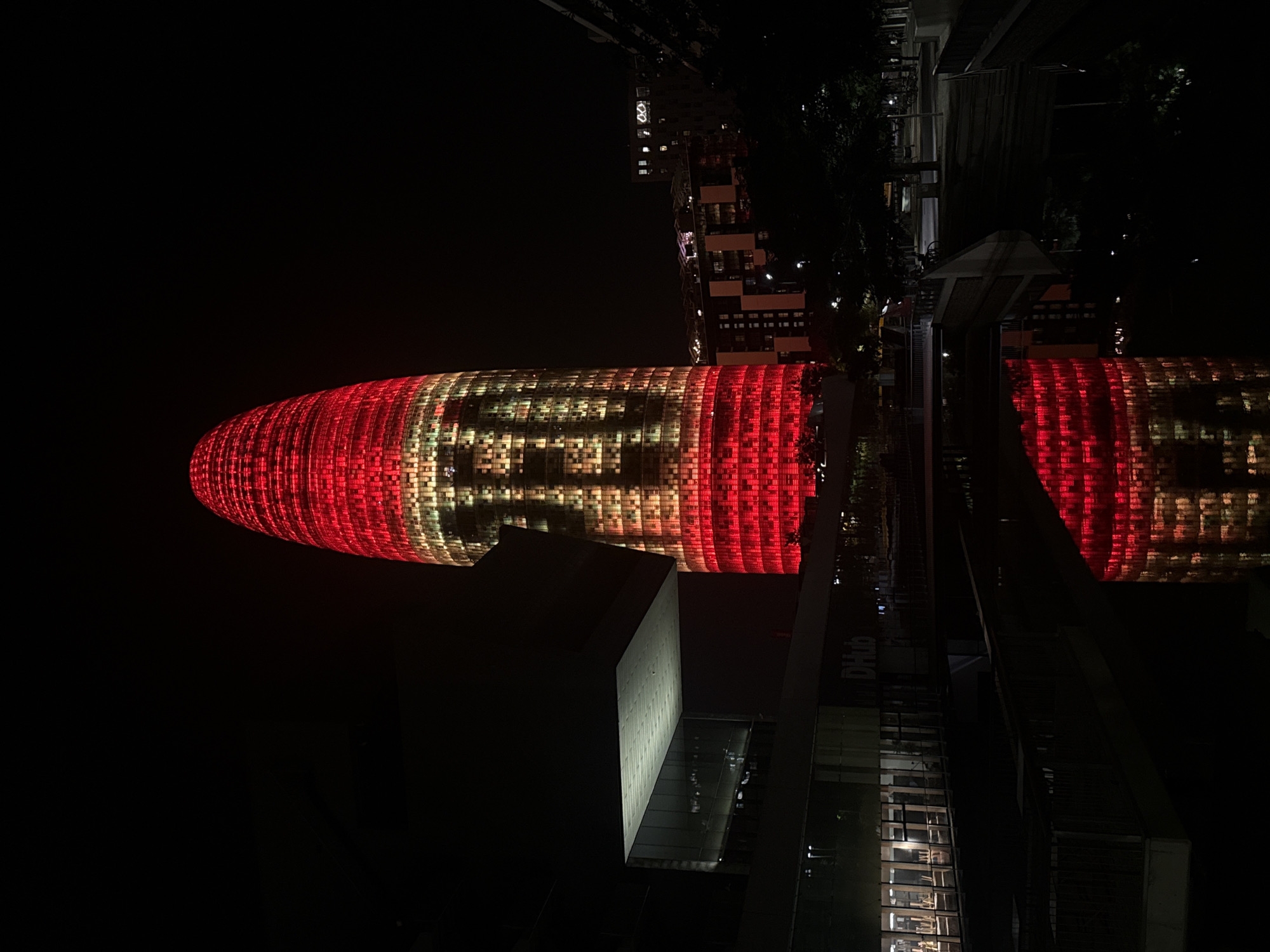 The image captures the Agbar Tower, a distinctive skyscraper in Barcelona, Spain, illuminated at night. The tower is predominantly lit in red, with sections of yellow and white light creating a pattern on its facade. The light display on the tower appears to form the numbers "100".
In the foreground, a body of water reflects the illuminated tower and the surrounding buildings. To the left of the tower, a modern building with a cantilevered section is visible, also lit from within. The surrounding environment is dark, typical of a nighttime urban scene, with trees and other structures partially visible. The scene conveys a festive or celebratory atmosphere due to the vibrant lighting of the iconic tower.