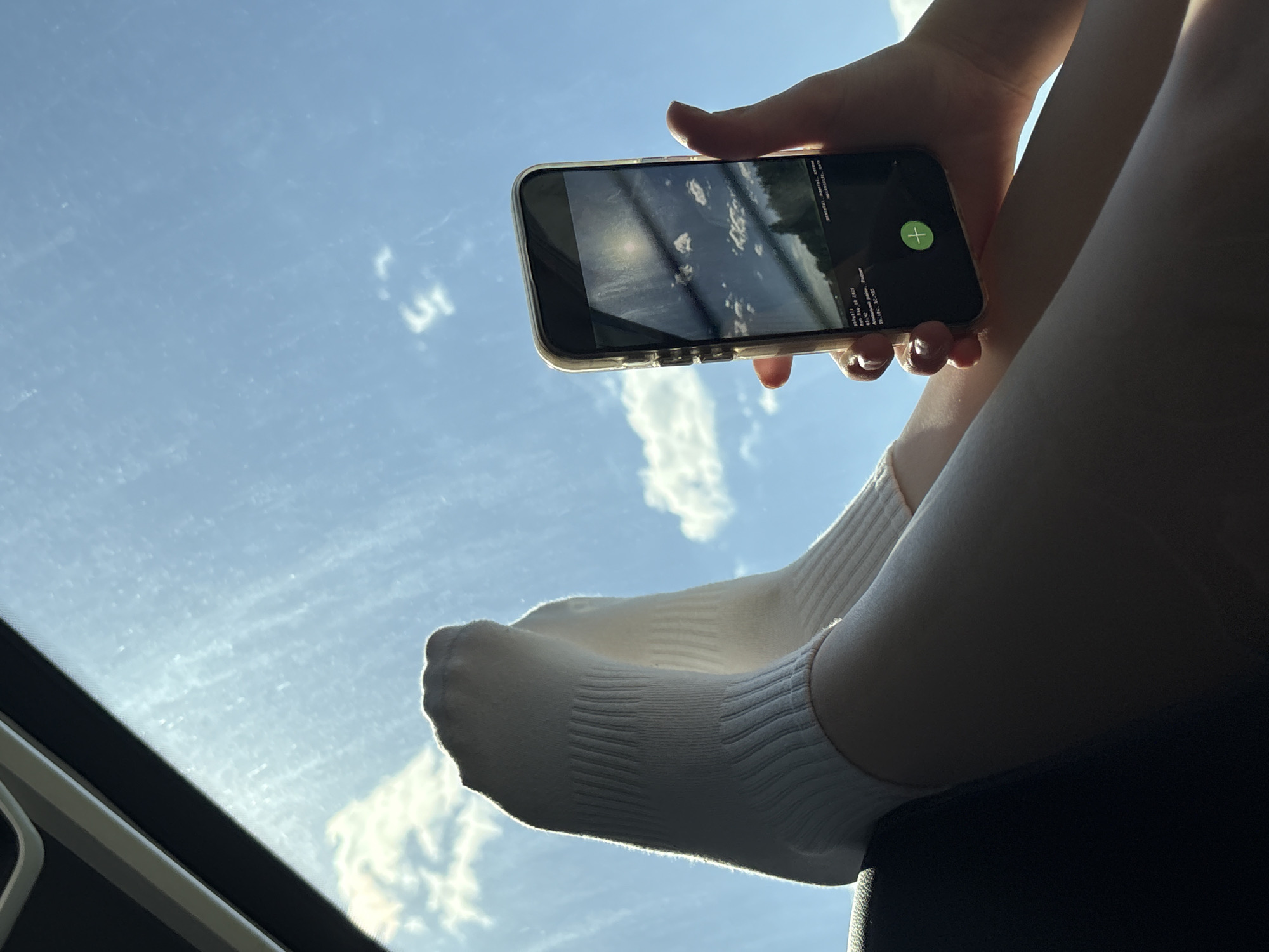 The photograph shows a person holding a smartphone in their left hand. The phone is displaying an image that appears to be a screenshot of the sky. The person is wearing white socks and is sitting inside what appears to be a vehicle with a sunroof. The sky visible through the sunroof is blue with scattered white clouds. Based on the text visible on the smartphone screen, the photograph was taken in Alnashi, Russia.
