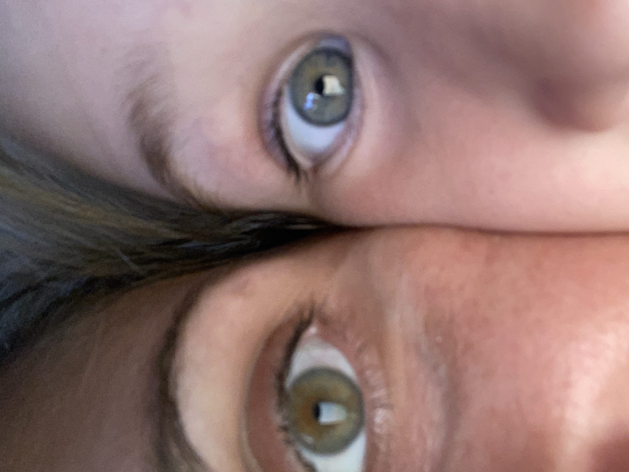 The image is a close-up, split view of two people's eyes, positioned side-by-side. The left eye, belonging to one individual, is a hazel color with visible iris detail and appears to be from an adult. The right eye, belonging to another individual, is a grey-blue color and appears to be from a child. The background of the image is blurred, with hints of hair and skin texture. There is no identifiable context of a city or specific location within Zgrade, Montenegro.