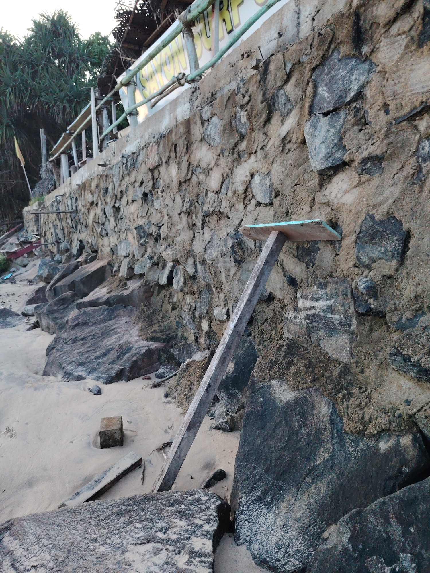 The image shows a stone wall on a beach. The wall is made of rough-hewn stones and mortar, and it appears to be supporting a walkway or railing above. A wooden plank with a light blue surface is propped against the wall, supported by a wooden beam. The beach itself is sandy, with several large rocks scattered about. Some debris, including what looks like broken wood and possibly plastic, is visible in the sand near the wall. Vegetation, likely palm trees, can be seen in the background to the left of the wall. The sky is overcast, suggesting a cloudy day. The lighting is soft, indicating it might be morning or late afternoon. There is some text on a banner or sign in the upper portion of the image, but it is partially obscured and difficult to read clearly. The overall scene suggests a somewhat neglected or rustic coastal area.