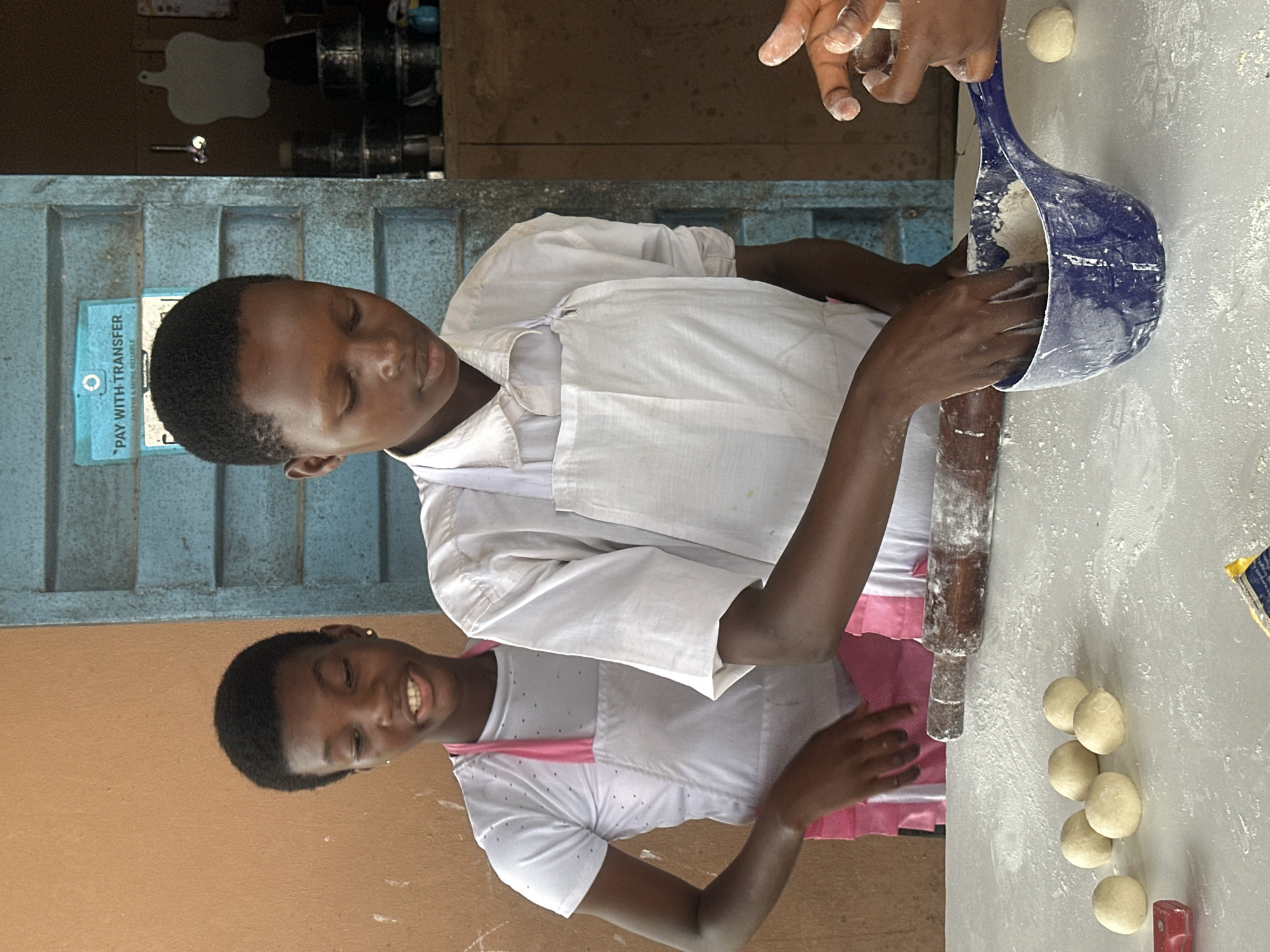 The image captures a candid moment of two young individuals, likely teenagers, engaged in a culinary activity in Badagry, Nigeria. The scene unfolds in what appears to be an indoor, possibly a community kitchen or a school setting, given the rustic wooden door with faded blue paint and a "Pay with Transfer" sticker.
The foreground is dominated by a white, flour-dusted surface, where several round dough balls are neatly arranged. A wooden rolling pin lies horizontally, also dusted with flour, suggesting its recent use. To the right, a blue plastic container, its rim and interior coated with flour, is being handled by a pair of hands. These hands, belonging to the individual in the white apron, are actively working with the flour inside the container.
The two main subjects are positioned behind the table. The individual on the right, facing slightly away from the camera, is wearing a white collared shirt and a white apron. Their gaze is directed downwards towards their hands and the blue container. The individual on the left, who is looking towards the camera with a slight smile, is wearing a white t-shirt with a subtle polka dot pattern, a pink apron, and a pink sash. Their expression suggests a cheerful engagement in the activity. Another pair of hands is visible in the lower right corner, also interacting with the flour and dough.
The background reveals a textured, ochre-colored wall on the left, contrasting with the blue door. Behind the door, shelves are visible, holding various containers and what appear to be bottles, hinting at stored ingredients or kitchen supplies. The overall lighting is natural, casting soft shadows and highlighting the flour-covered surfaces. The scene is one of communal activity and learning, evoking a sense of productivity and youthful engagement in a practical skill.