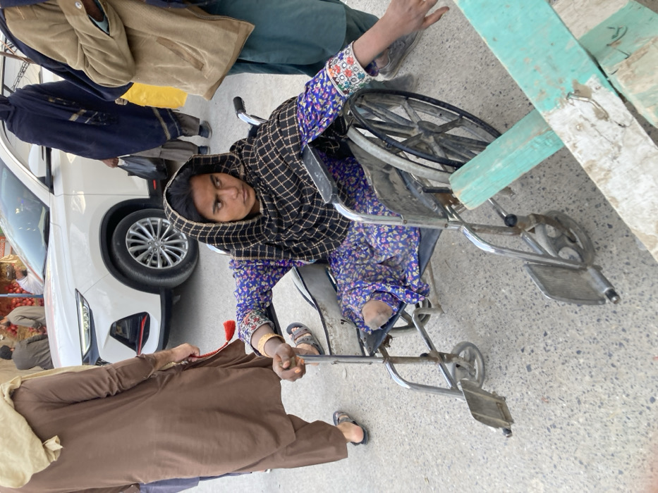 In Quetta City Tehsil, Pakistan, a street scene captures a young woman seated in a manual wheelchair. The woman wears a purple floral patterned garment, a dark checkered headscarf, and has gold-colored bangles on her right wrist, with colorful embroidery on her left sleeve. She is missing both legs above the knee, with the stumps visible. Her right hand rests on the wheelchair armrest, while her left arm extends forward. In the mid-ground, a man in a brown shalwar kameez and light head covering walks to the left. To the right, another man partially visible wears a tan top and green garment. A white SUV is parked behind the woman. Further in the background, a woman in dark attire walks away from the camera, carrying a black bag, and two men stand near a fruit stall displaying red pomegranates. The ground is a paved surface, and rough, light blue/green wooden planks are present in the immediate foreground, partially obscuring the view.