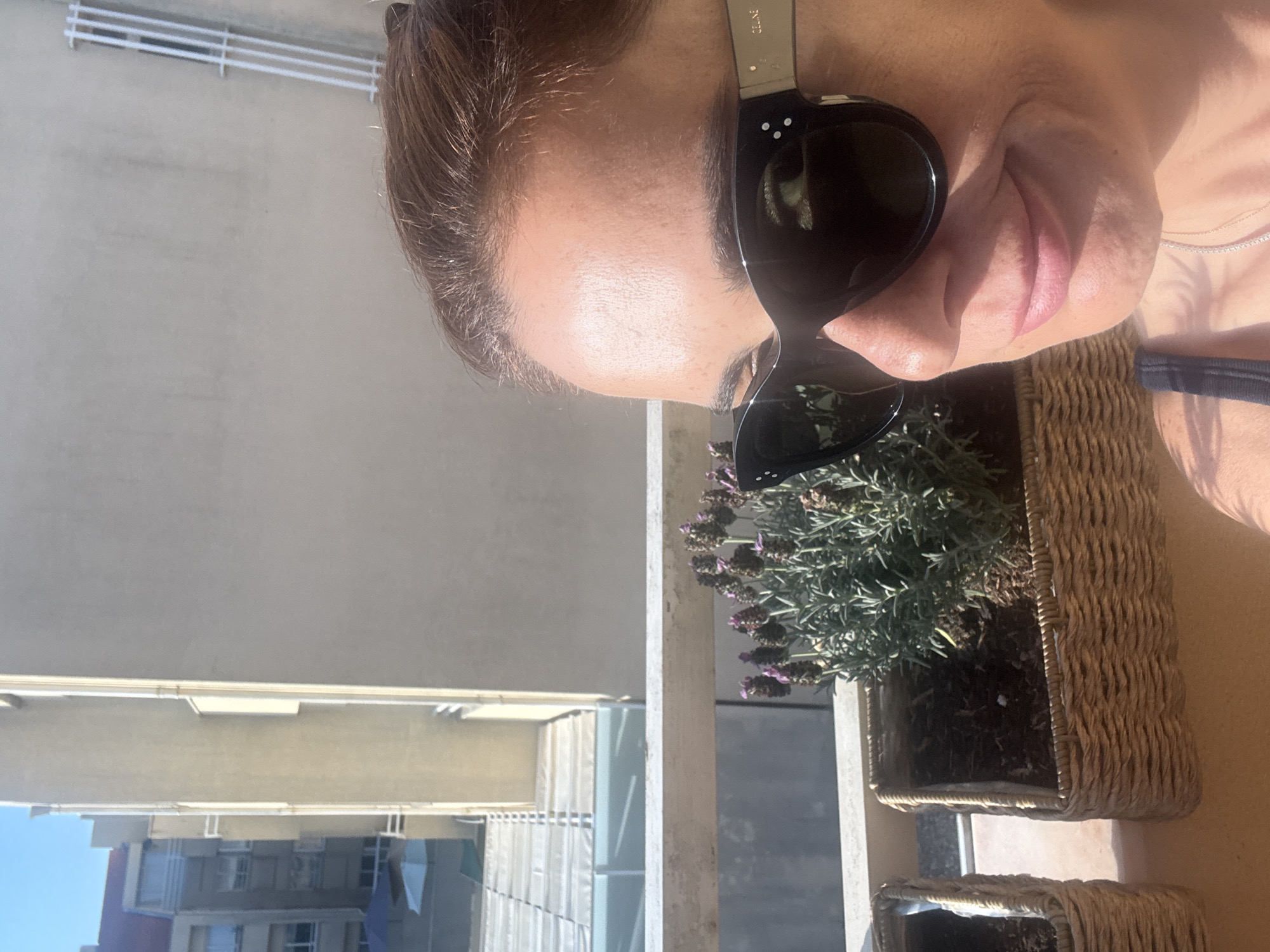 In Vila Nova de Gaia, Portugal, a woman is pictured on a balcony, her face close to the camera, wearing large, dark sunglasses. Behind her, a building with a light beige facade dominates the background, with parts of another building visible to the left. On the balcony railing, a planter box filled with soil holds a plant with delicate purple flowers, possibly lavender. Another planter box, partially visible to the left, appears empty or contains dark soil. The sunlight casts a warm glow on the scene, illuminating the woman's skin and the textures of the planter boxes. The woman is wearing a dark-colored top, and a delicate necklace is visible around her neck. The image appears to be a casual selfie taken outdoors on a sunny day.